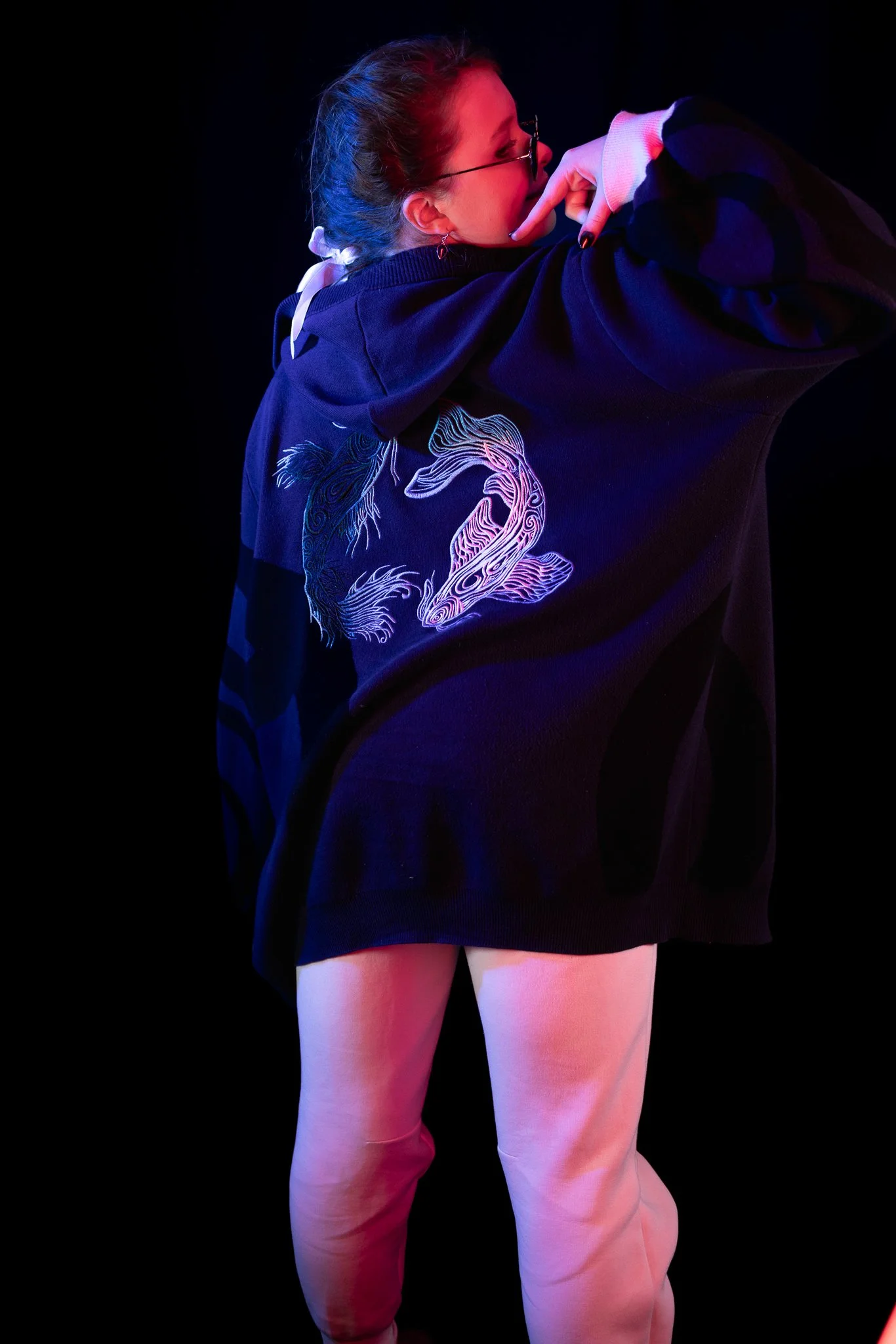 A woman with glasses and earrings is posing in a dark setting with colorful neon lighting. She is wearing an oversized black hoodie with a glowing fish design on the back and light-colored pants.
