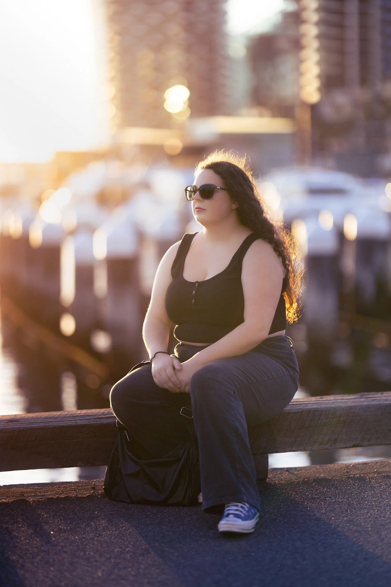 A woman wearing sunglasses and a black crop top with jeans, sitting on a wooden barrier by the water during sunset, with boats and buildings in the background.