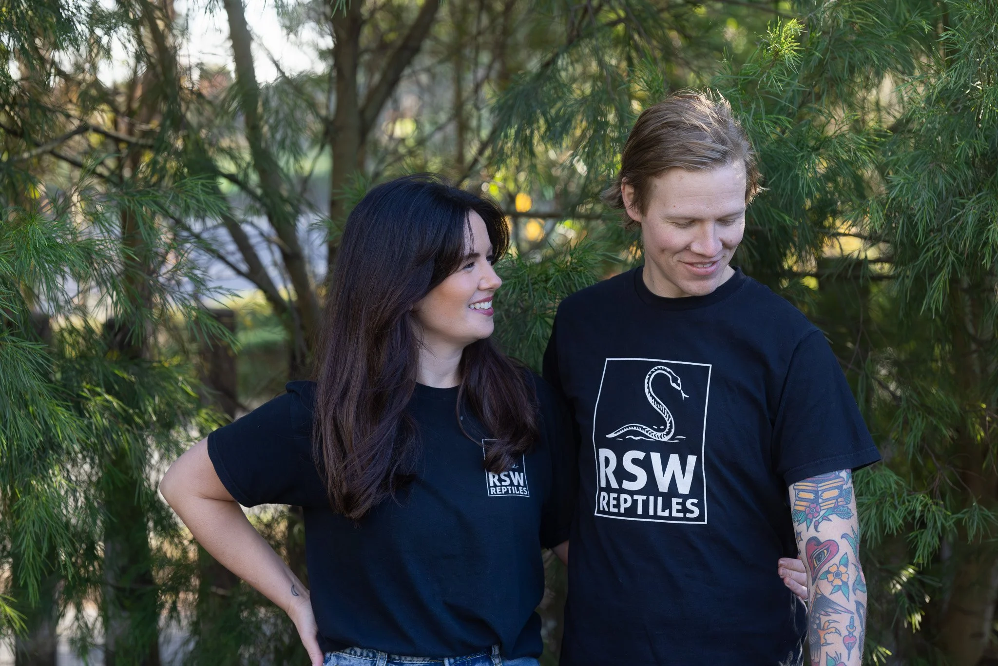 Two people standing outdoors in front of green trees, wearing black T-shirts with the 'RSW REPTILES' logo, smiling and looking at each other.