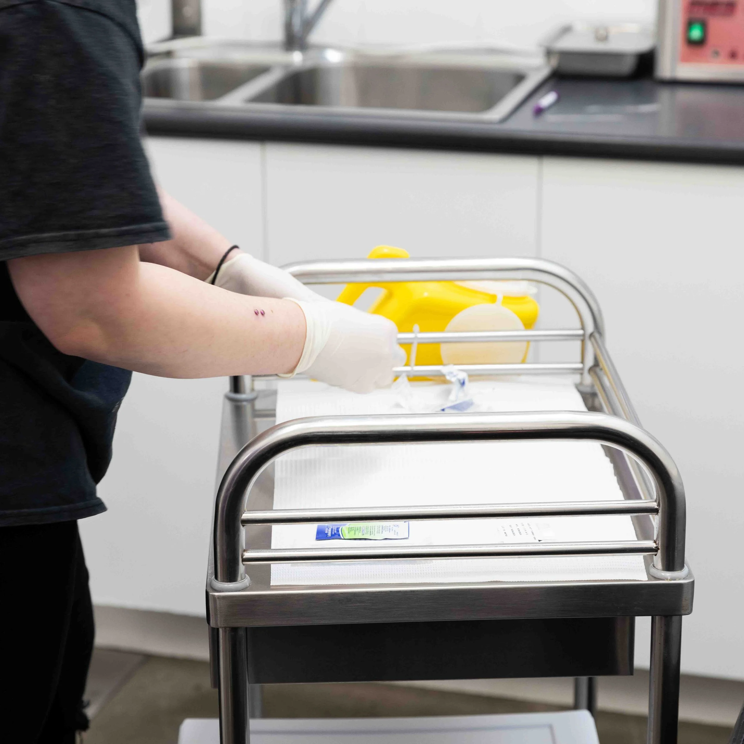 Healthcare worker wearing gloves preparing medical supplies on a stainless steel cart in a clinical setting.