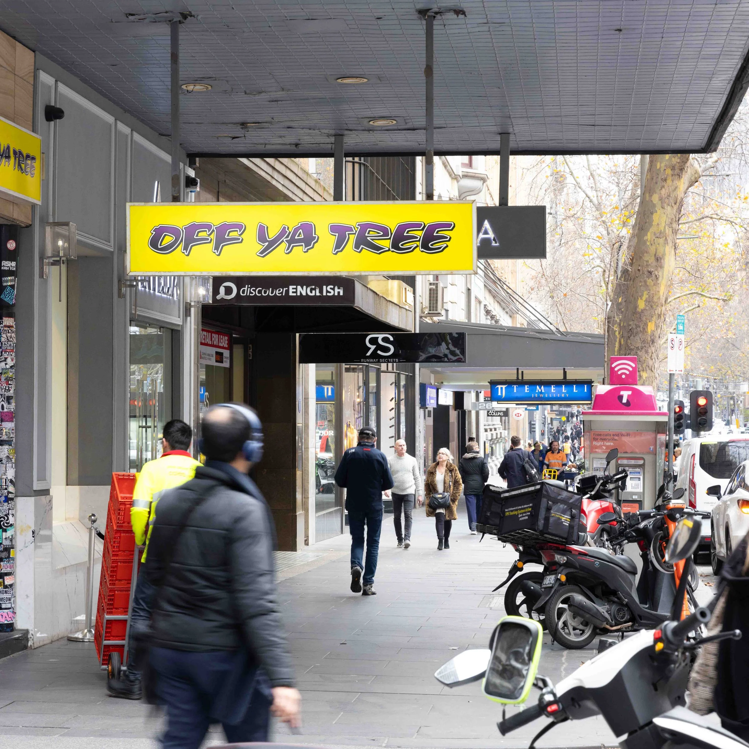 City sidewalk scene with people walking past storefronts, including a bright yellow sign that reads 'OFF YA TREE' and various shops.