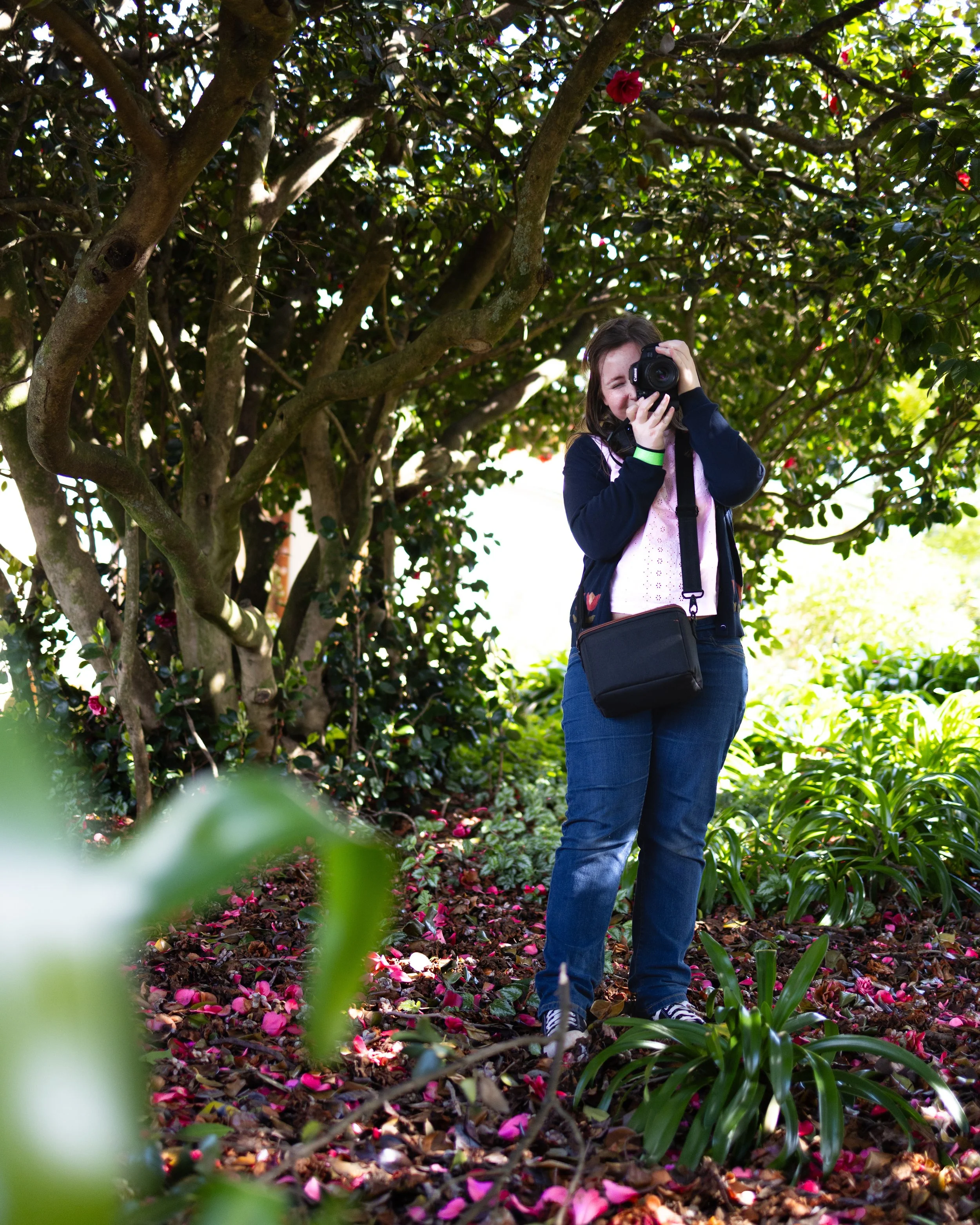 A woman standing in a garden surrounded by trees and plants, taking a photograph with a camera.