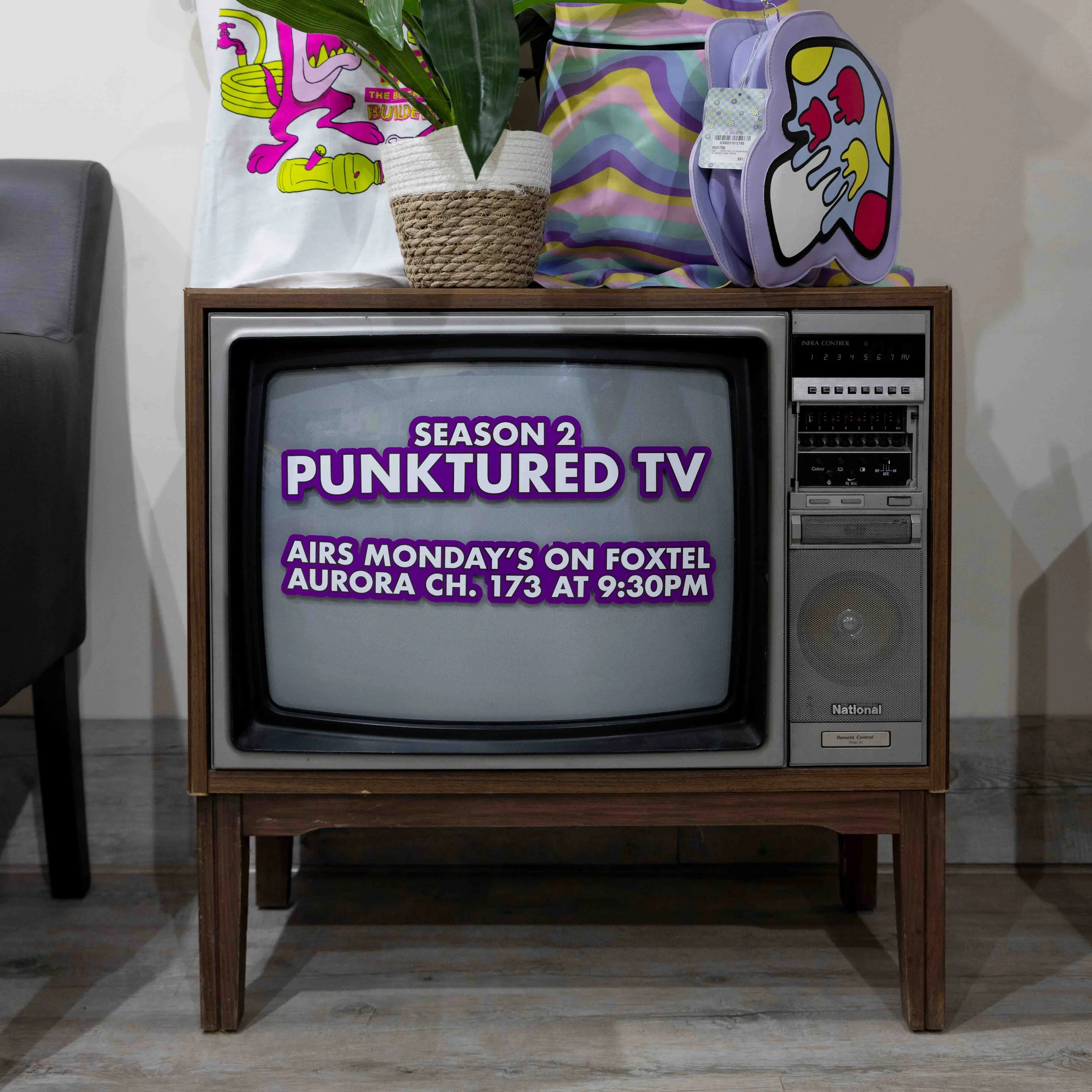An old-fashioned wooden TV set displaying a program called 'Punkatured TV' with purple text, along with details for a show airing on Foxtel at 9:30 PM. The TV has various items on top, including a potted plant, a colorful bag, and a tote bag with a p