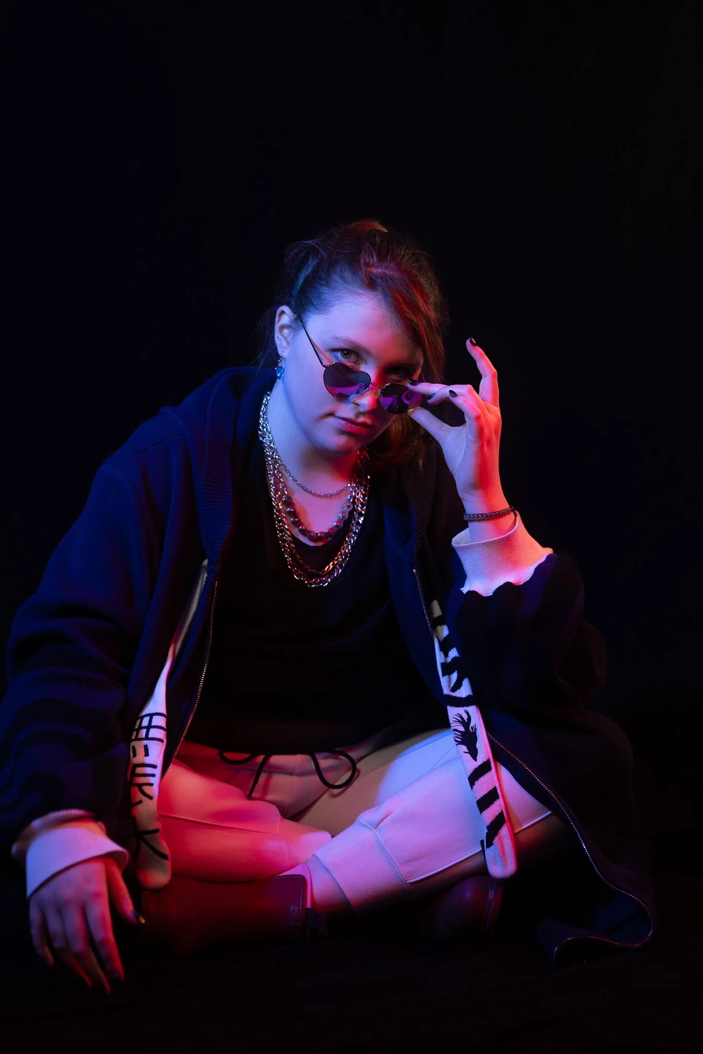 A young woman sitting cross-legged against a black background, wearing sunglasses, layered necklaces, a hoodie, and beige pants, illuminated with colorful neon lighting.