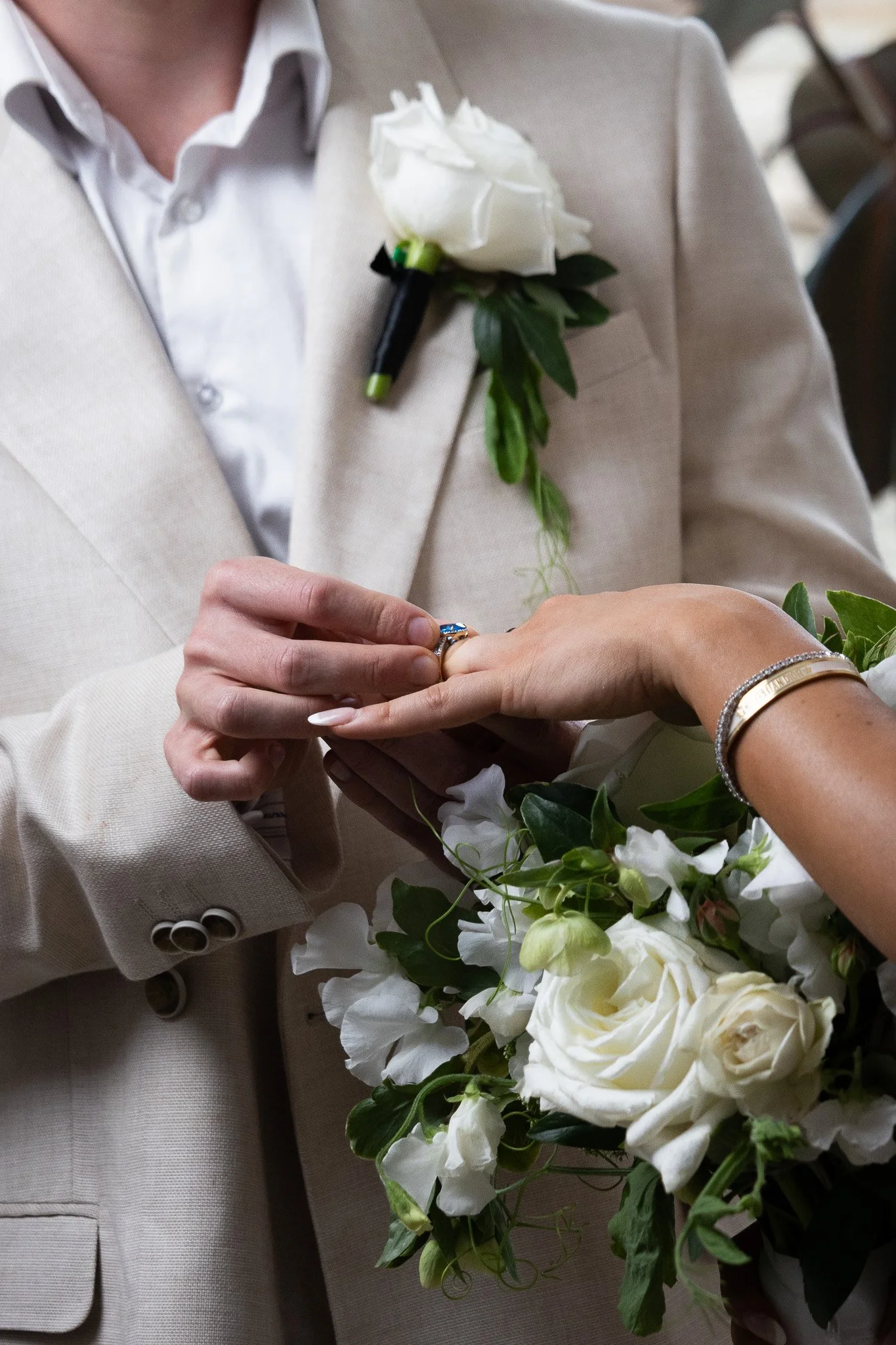A man in a beige suit and white shirt is holding a woman's hand, as she is placing a ring on his finger during a wedding ceremony. The woman is wearing a silver bracelet, and there is a bouquet of white flowers with green leaves in the foreground.