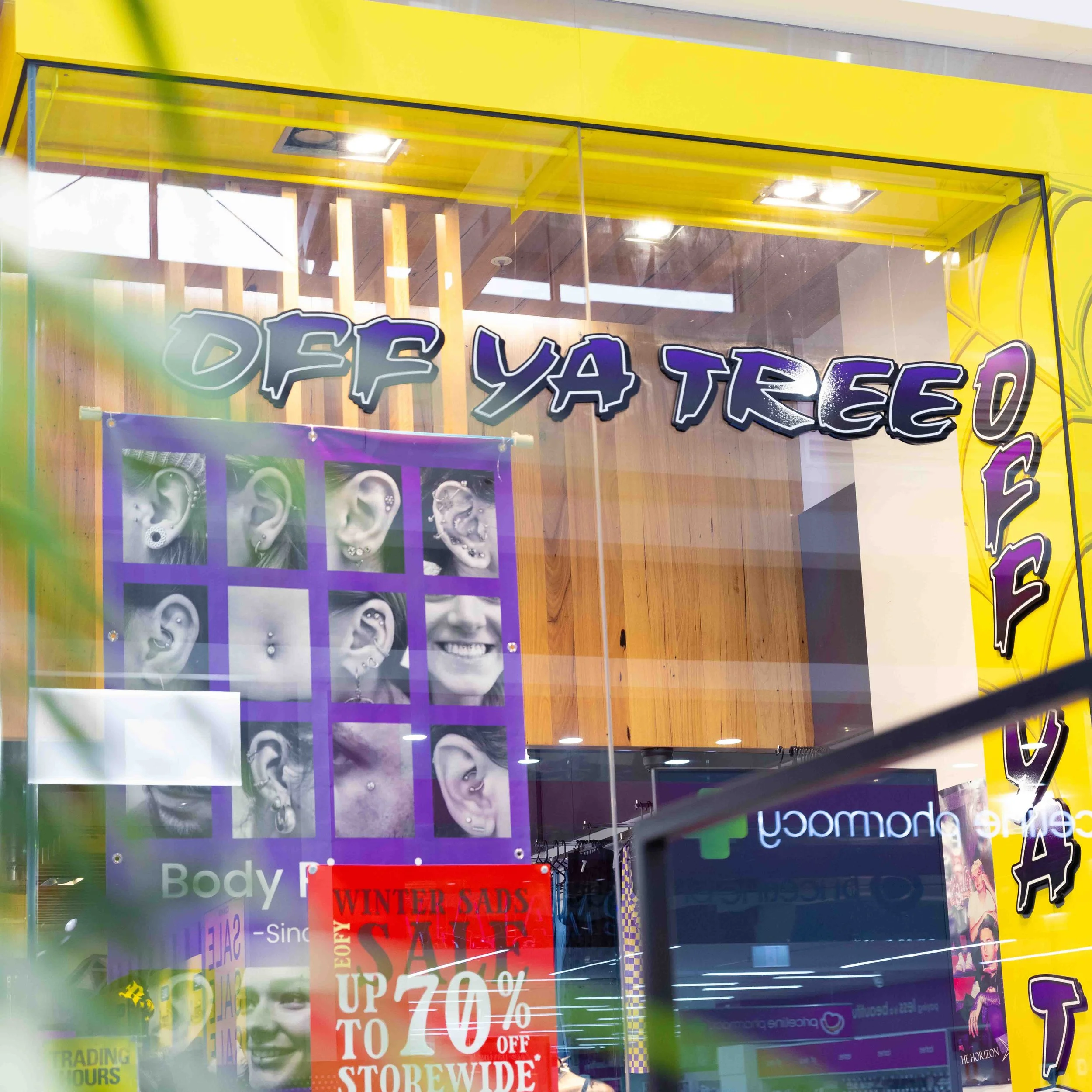 Storefront window with signage and posters advertising body jewelry, a sale of up to 70% off, and displaying photos of various piercings.