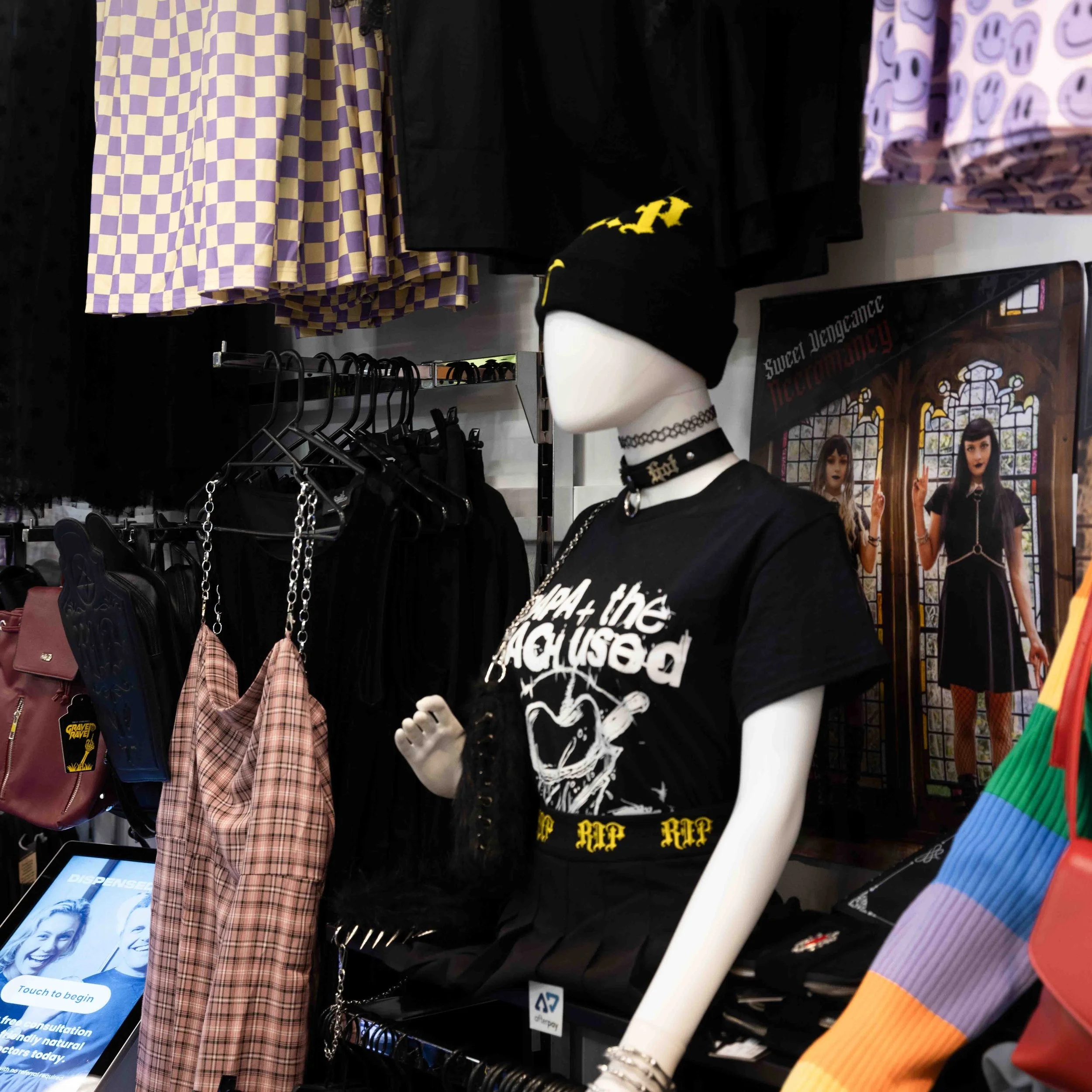 A clothing store display featuring a mannequin dressed in goth-inspired attire, including a black beanie, choker, graphic t-shirt, and black skirt. Behind the mannequin are racks of clothes, including checkered and dark-colored garments, and a poster