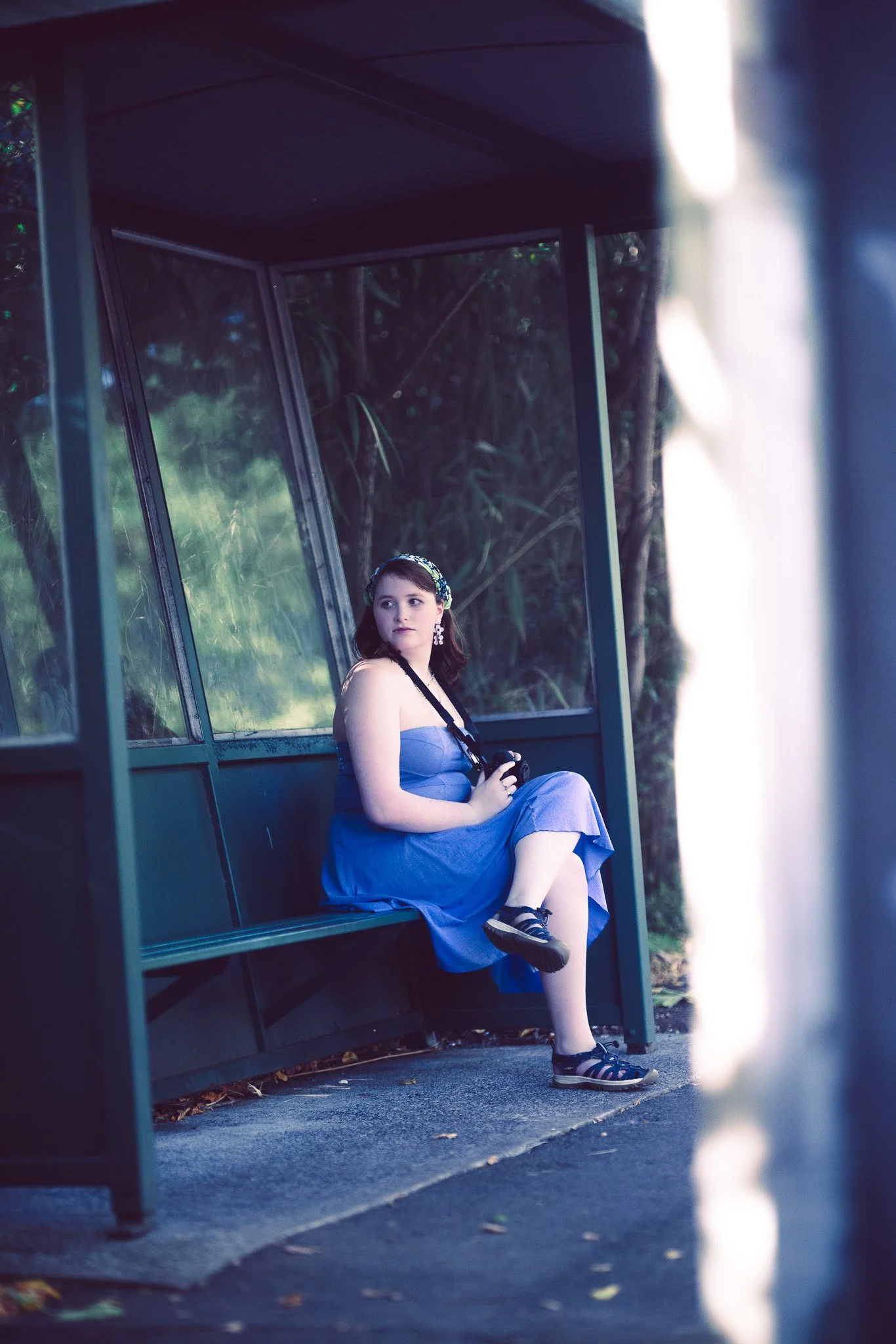 A young woman in a blue dress sitting on a bus stop bench, holding a camera, surrounded by greenery.