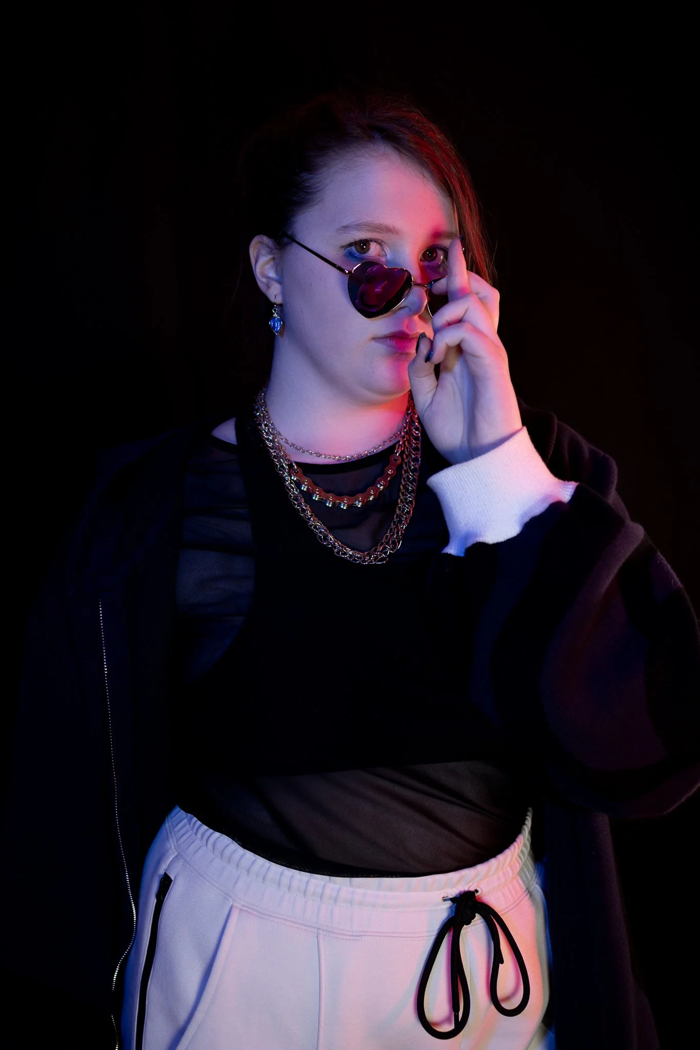 A young woman with dark hair and earrings wears layered chains and sunglasses, touching her face against a black background illuminated with colorful pink and blue lighting.