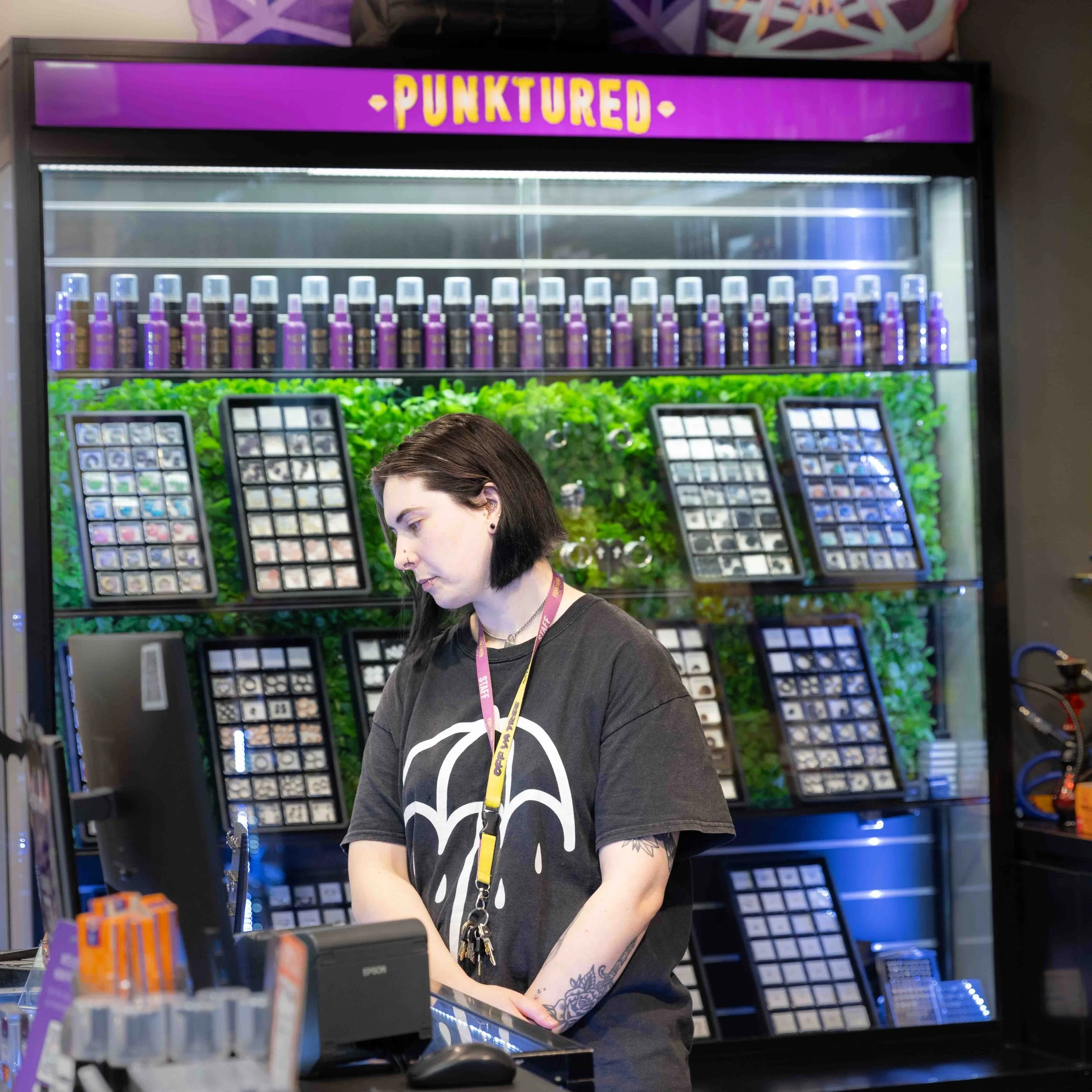 A young woman with dark hair and tattoos working at a retail counter, with a display of small bottles and framed items behind her, and a neon sign that says 'PUNKTURED' above.