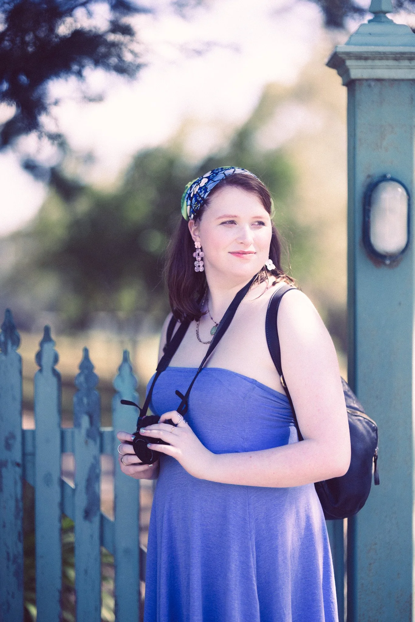 A young woman in a blue dress holds a camera, standing outdoors near a green fence and a vintage post box, with trees in the background.