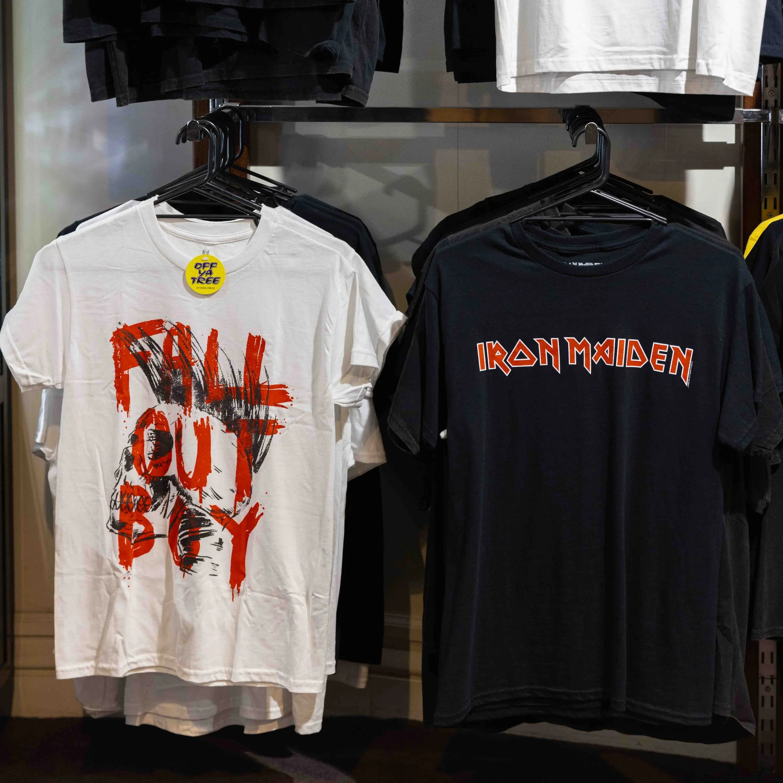 Two graphic T-shirts hanging on a clothing rack. The shirt on the left is white with a red and black skull design and the words "FALL OUT BOY" in red. The shirt on the right is black with the band name "IRON MAIDEN" in orange and white letters.