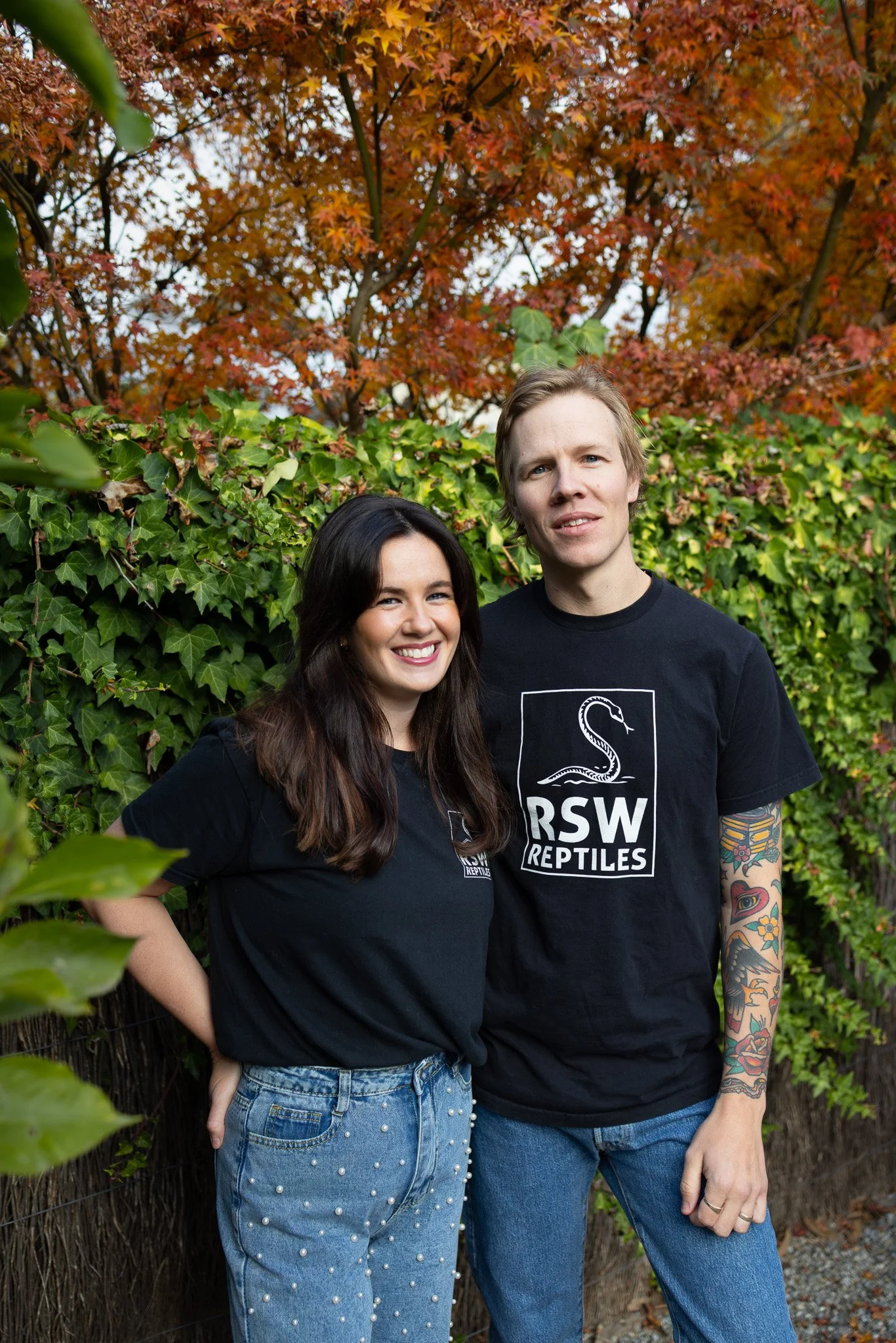 A smiling woman and a neutral man standing outdoors in front of green ivy and fall-colored tree leaves, both wearing black T-shirts with a snake and the words "RSW Reptiles".