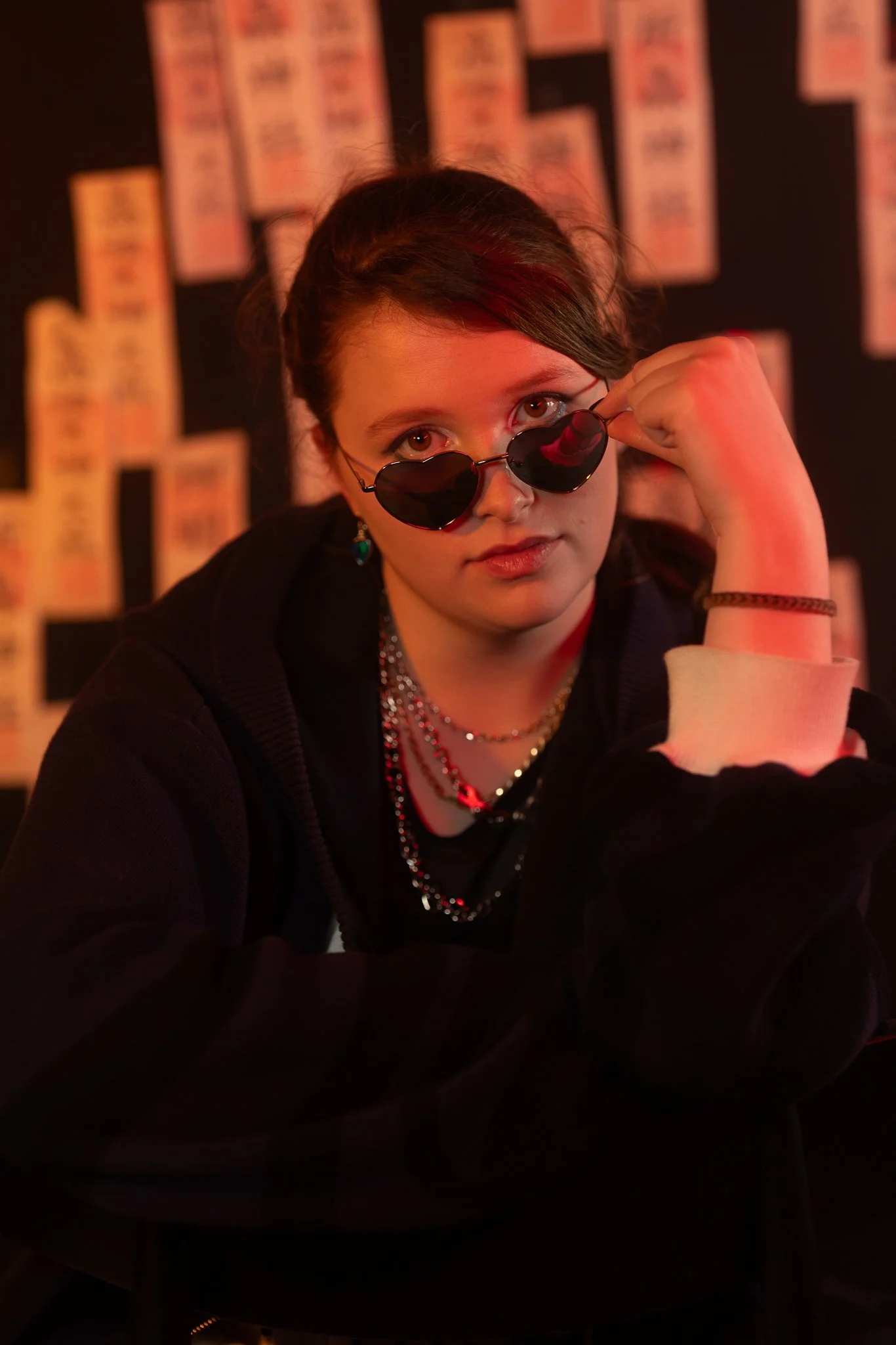 A young woman with dark hair, wearing heart-shaped sunglasses, layered necklaces, and earrings, sitting at a table with a dark background covered in colorful posters or prints, illuminated by red lighting.