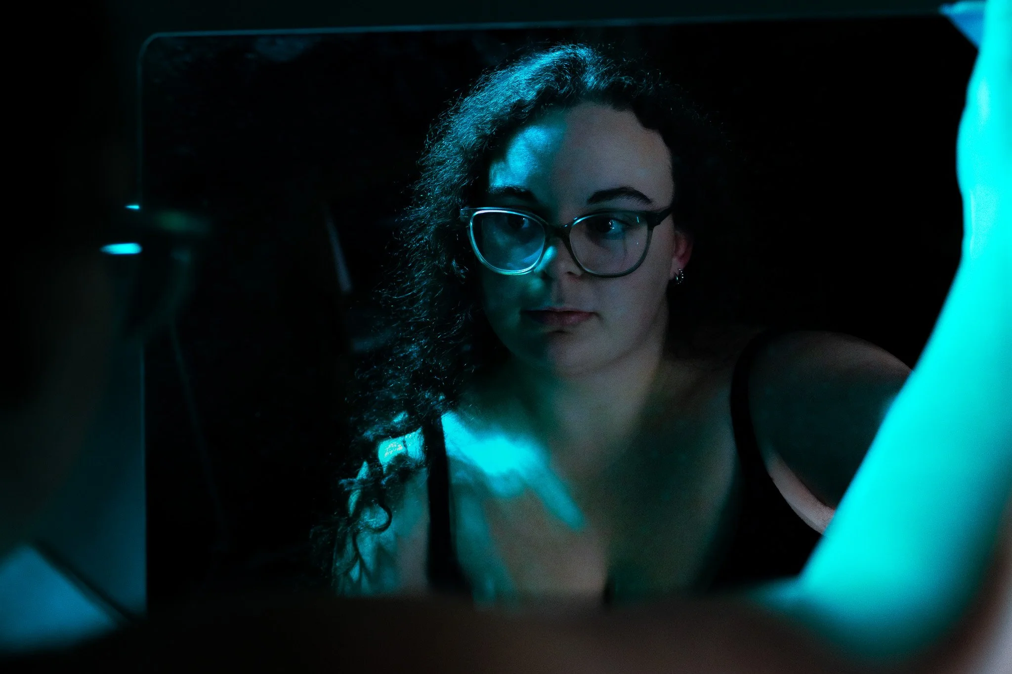 Woman with glasses looking at herself in a mirror illuminated with blue light.