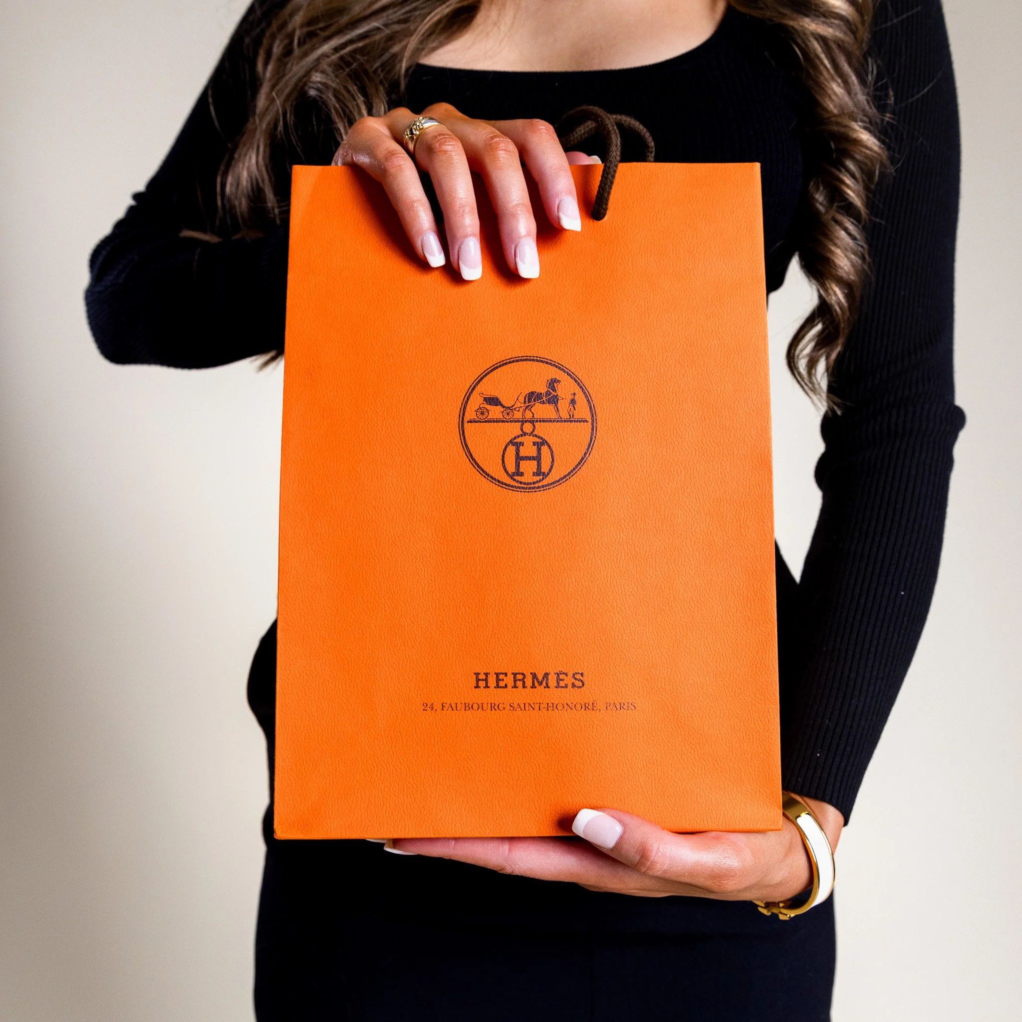 Person holding an orange Hermès shopping bag with black logo and text, wearing a black long sleeve top and jewelry.