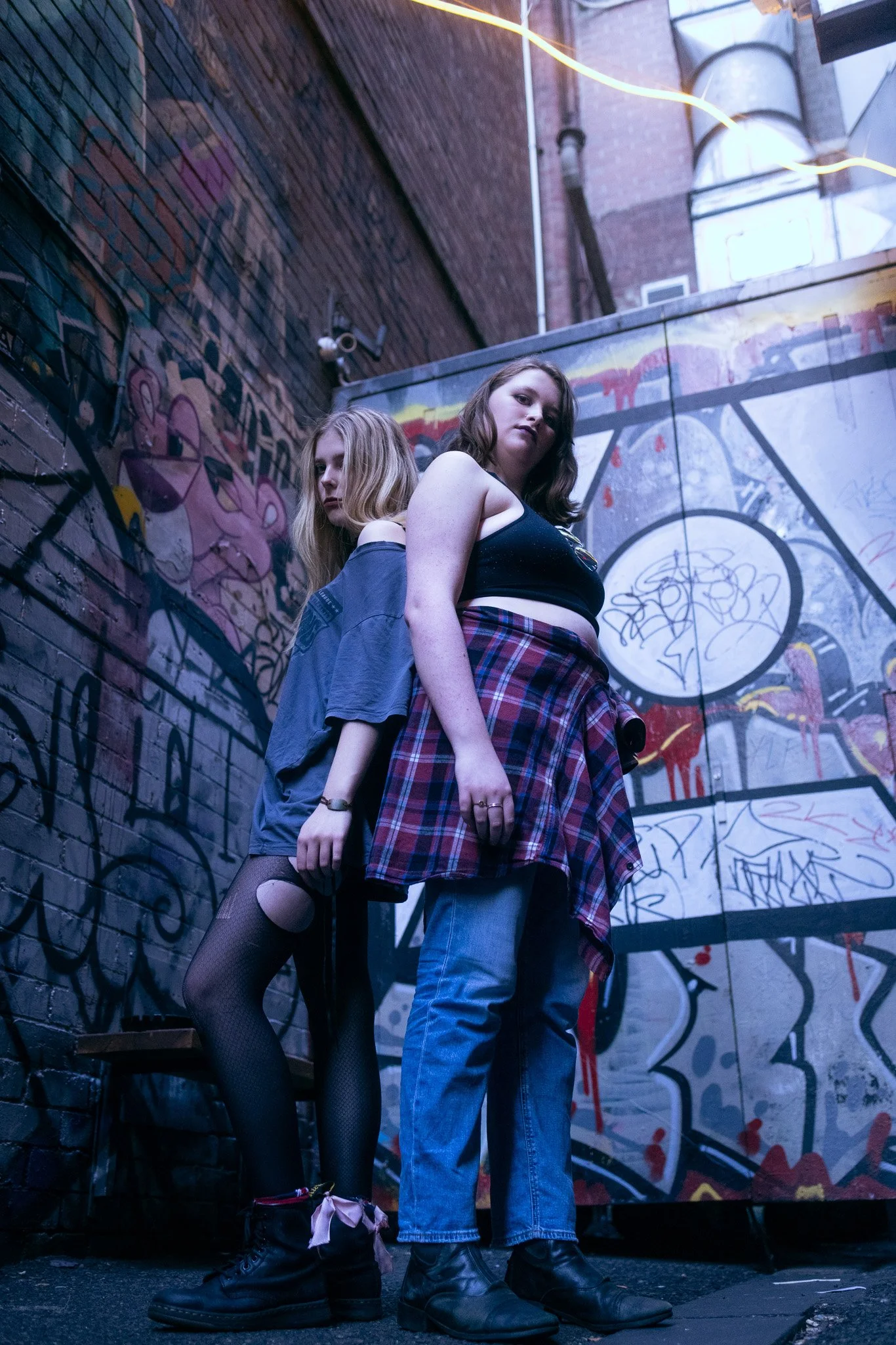 Two young women standing back-to-back in an alley with graffiti-covered walls, one wearing a black crop top and a plaid shirt tied around her waist, the other wearing an oversized blue shirt and ripped fishnet tights.