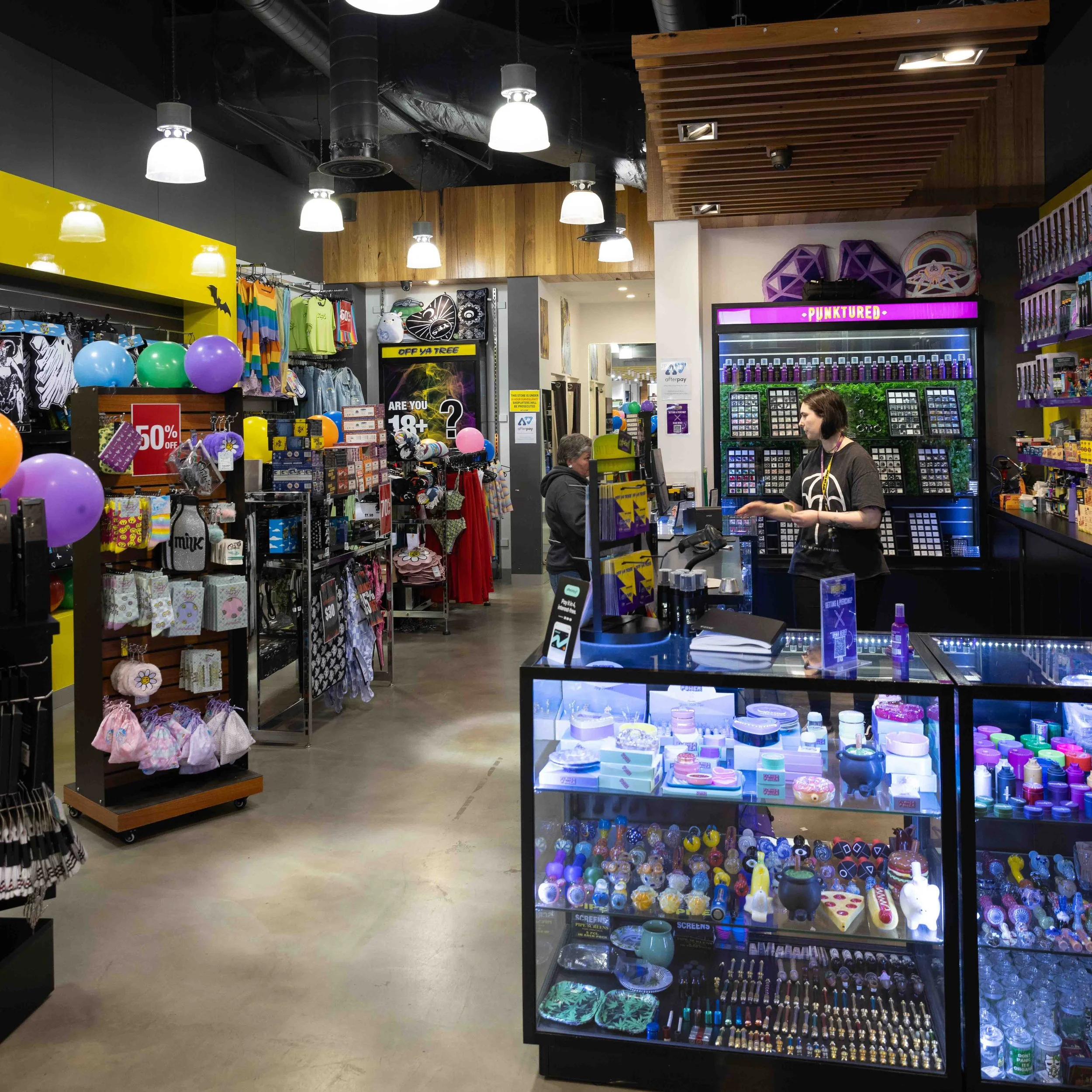Interior of a retail store with various products on display, including balloons, clothing, accessories, and vaping products, with a cashier and a customer at the checkout counter.
