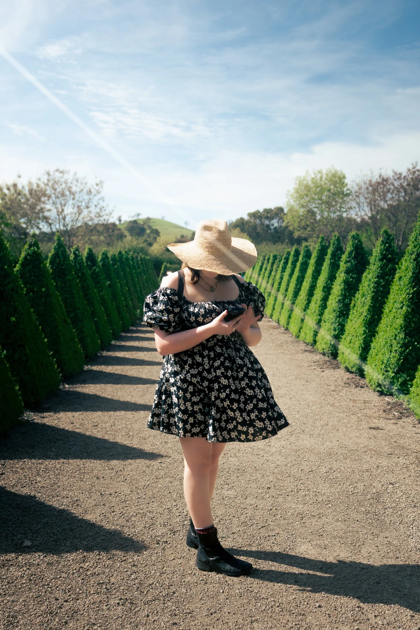A woman wearing a black floral dress, black boots, and a large straw hat stands in a vineyard, looking at her phone under a partly cloudy sky.