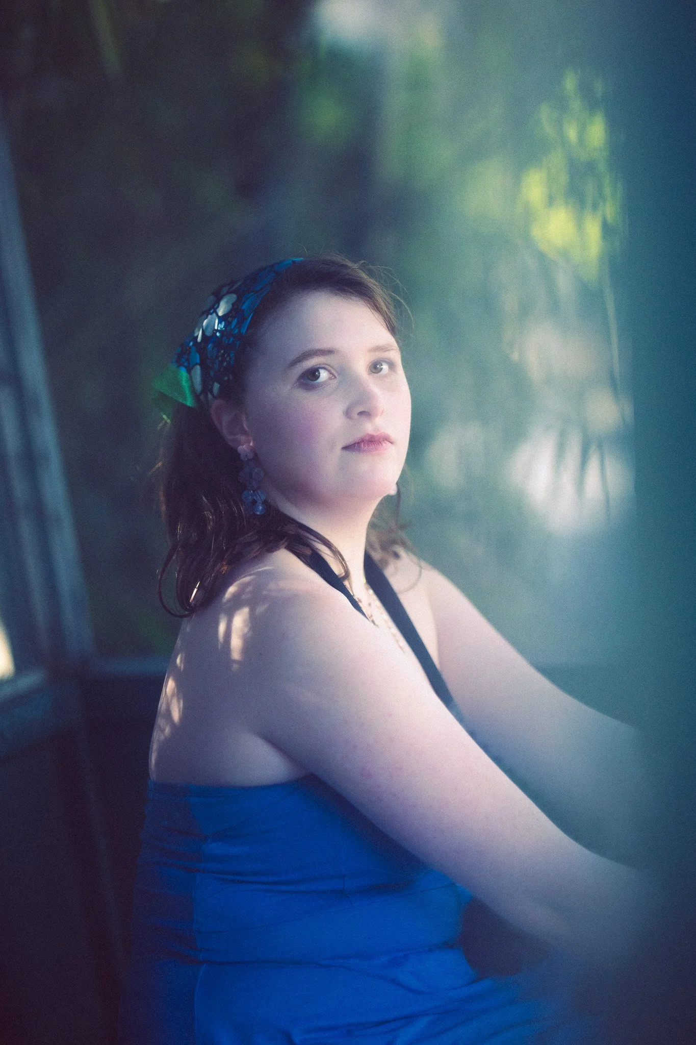 A woman with fair skin, brown hair, and blue eyes, wearing a blue dress, colorful earrings, and a headscarf, sitting by a window with a blurred outdoor background.
