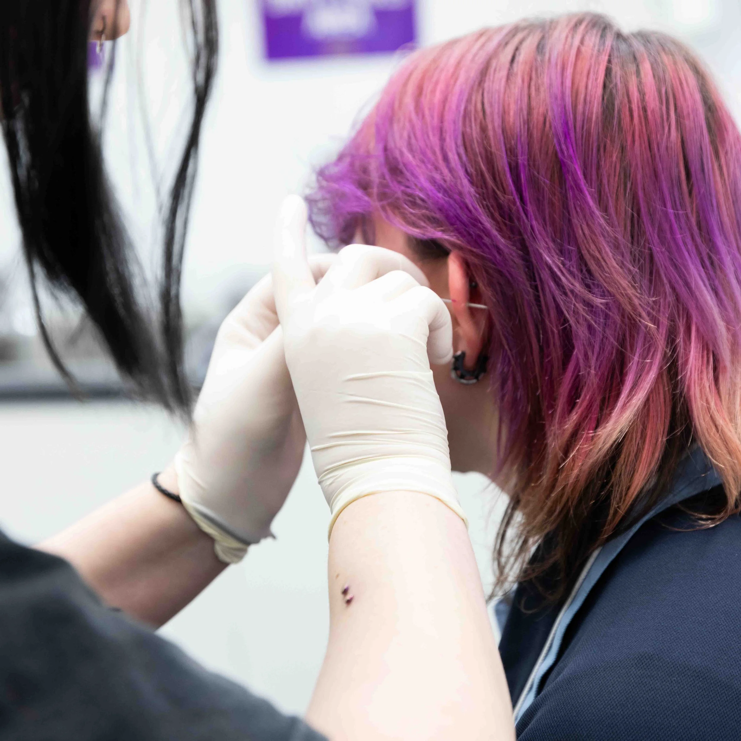 A person with pink and purple hair getting an ear piercing at a tattoo or piercing shop, with a professional wearing gloves administering the piercing.