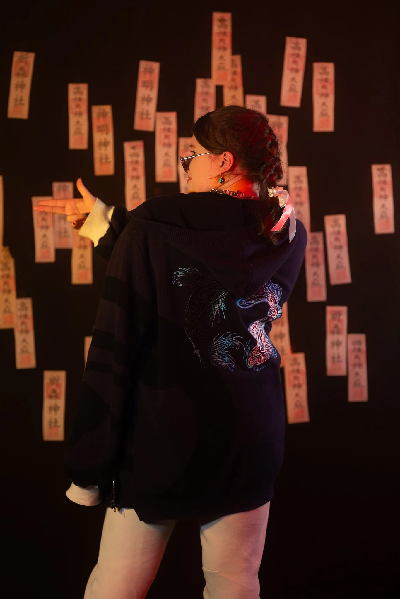 A woman with braided hair, sunglasses, and teal earrings points at a wall covered with pink paper strips with Japanese writing.