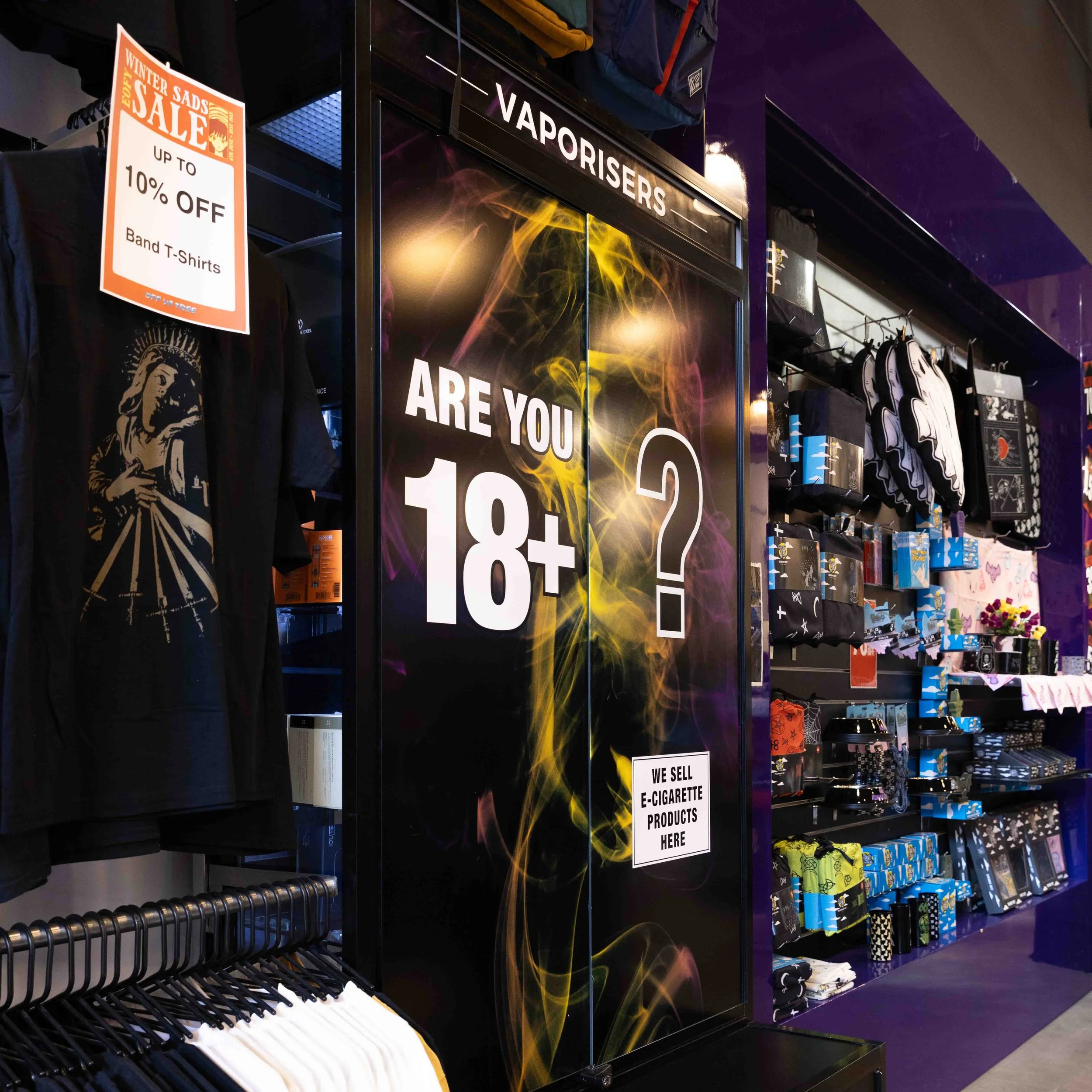 Store display with a sign asking, 'Are you 18+?', and shelves of sunglasses and accessories. An orange sale sign advertises up to 10% off band T-shirts.