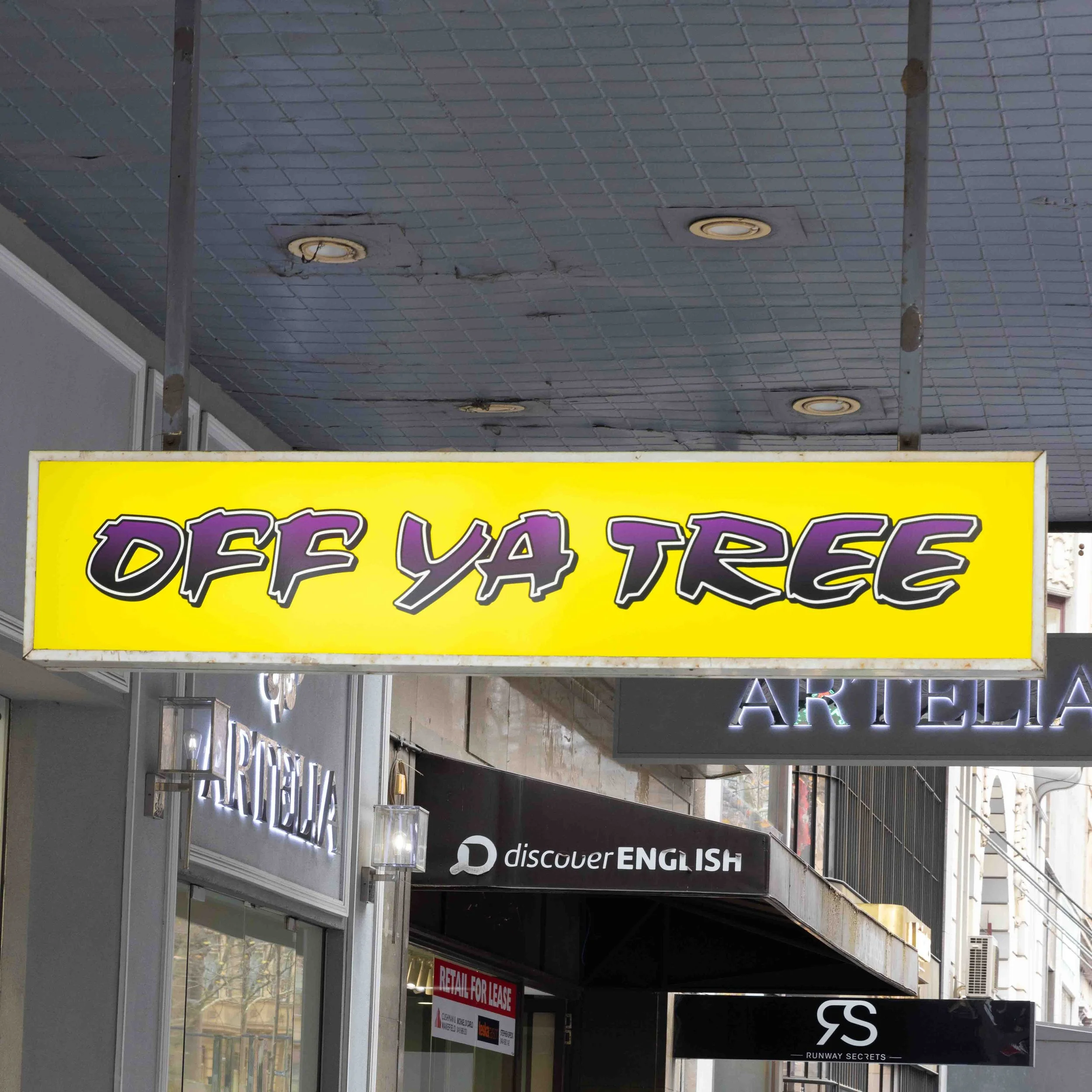 Bright yellow sign with purple and black text that reads 'OFF YA TREE' hanging outside storefronts in an urban shopping area.