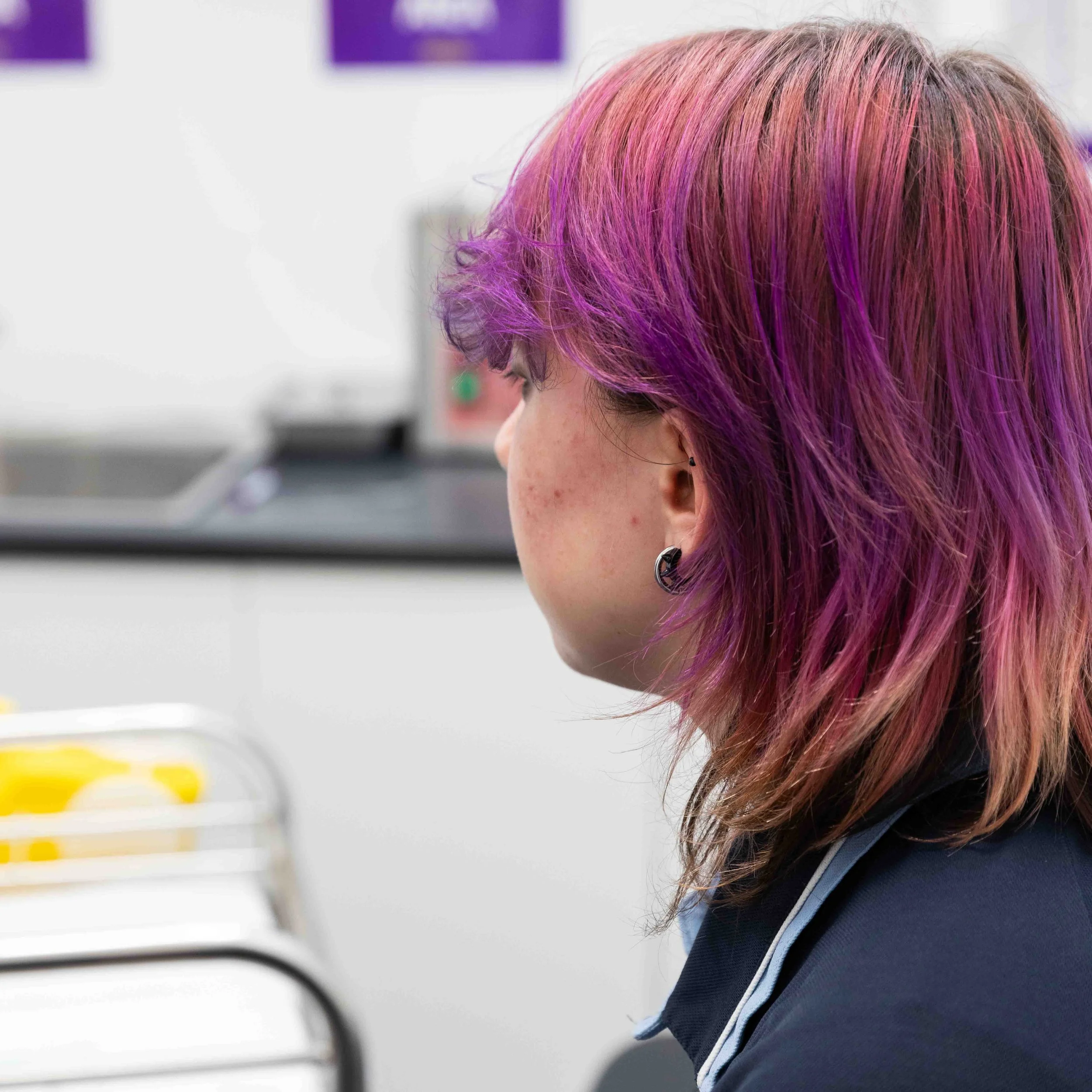 A person with shoulder-length hair dyed in shades of pink and purple, wearing an earring, sitting in an indoor setting.
