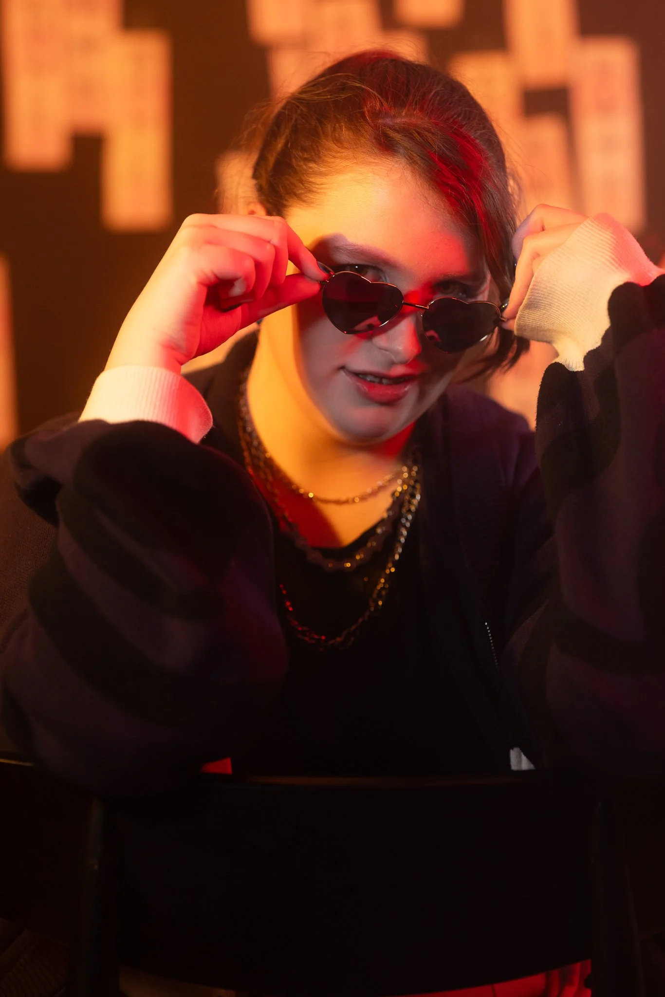 A person with brown hair wearing sunglasses, a black jacket, and layered necklaces, looks at the camera indoors with red and warm lighting.