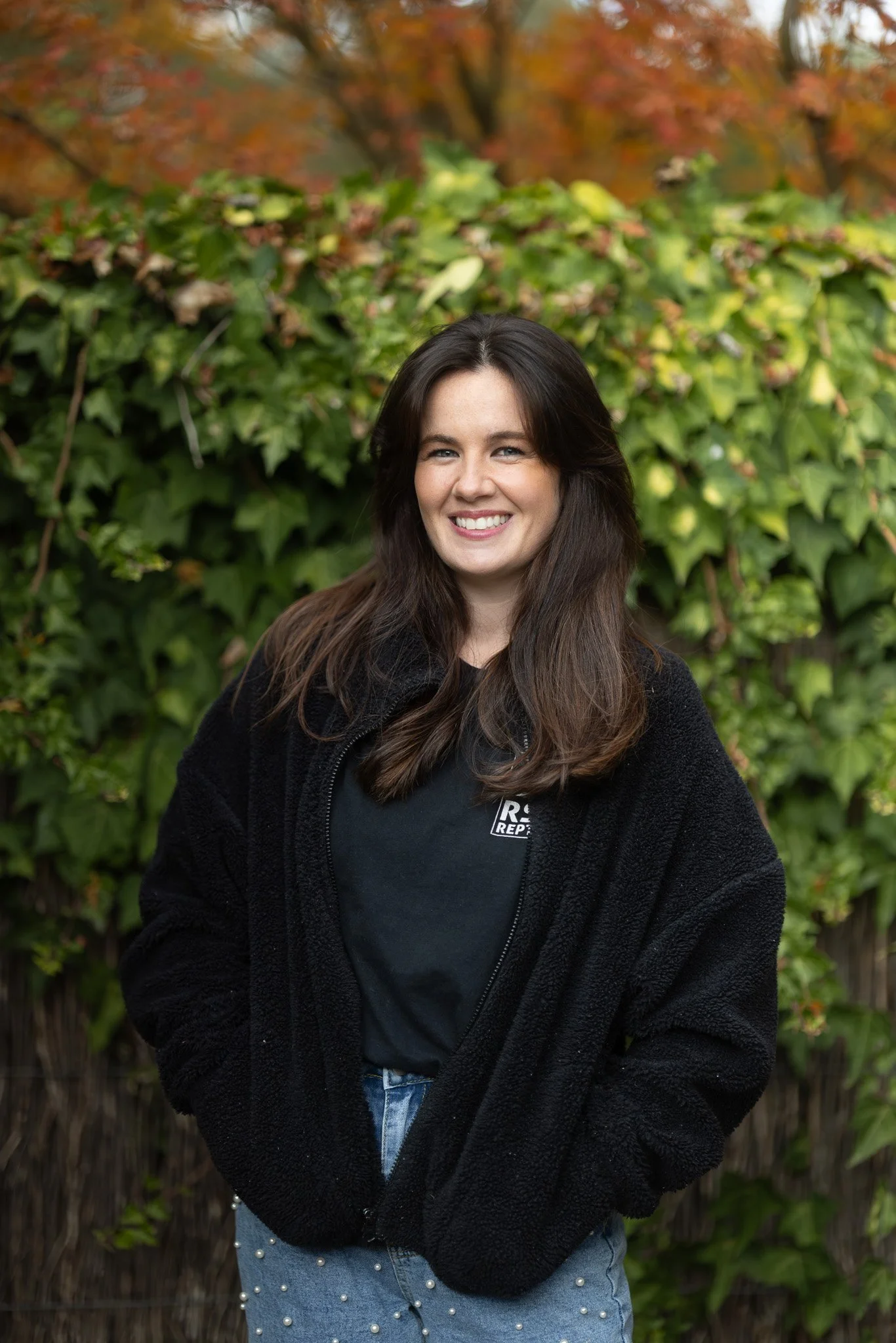 A young woman with long dark hair smiling outdoors in front of green ivy and autumn trees. She is wearing a black fleece jacket and a black t-shirt.