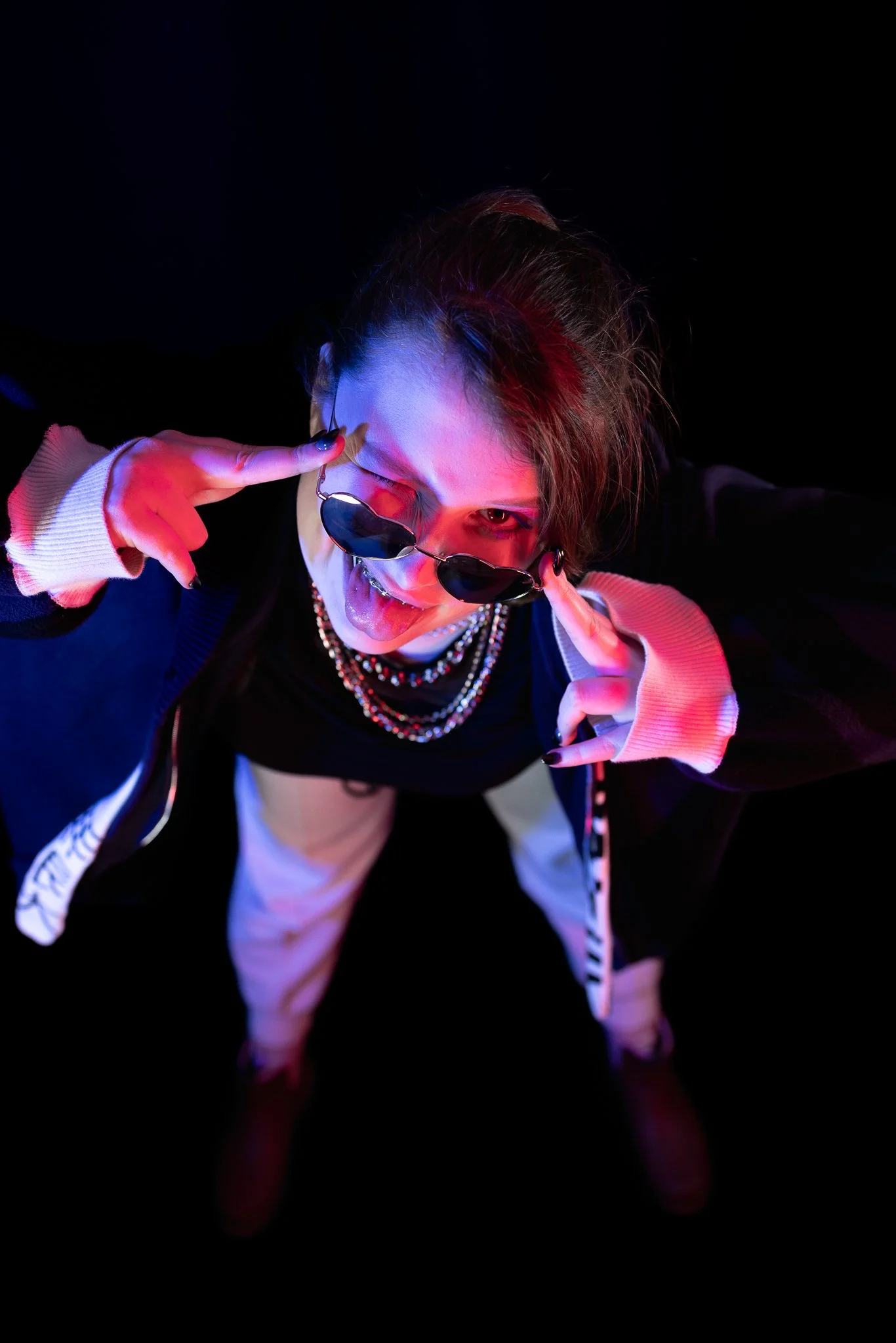A person wearing sunglasses, sticking out their tongue, and pointing to their temple with fingers, against a dark background, illuminated by colorful lighting.