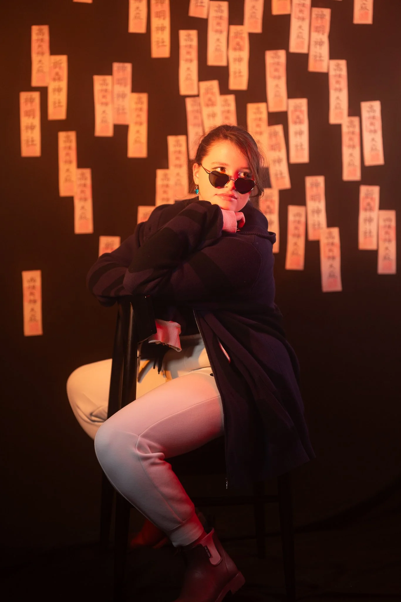 A woman with dark hair wearing sunglasses, a dark jacket, and beige pants sits on a black chair against a black wall decorated with numerous pieces of paper featuring Asian characters.