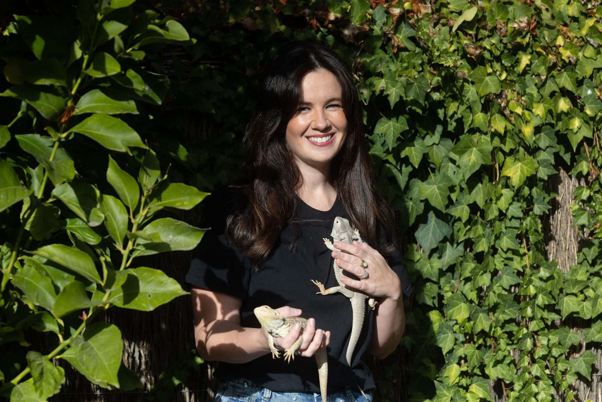 A woman with long dark hair smiling outdoors, holding two lizards in her hands against a background of green ivy leaves.