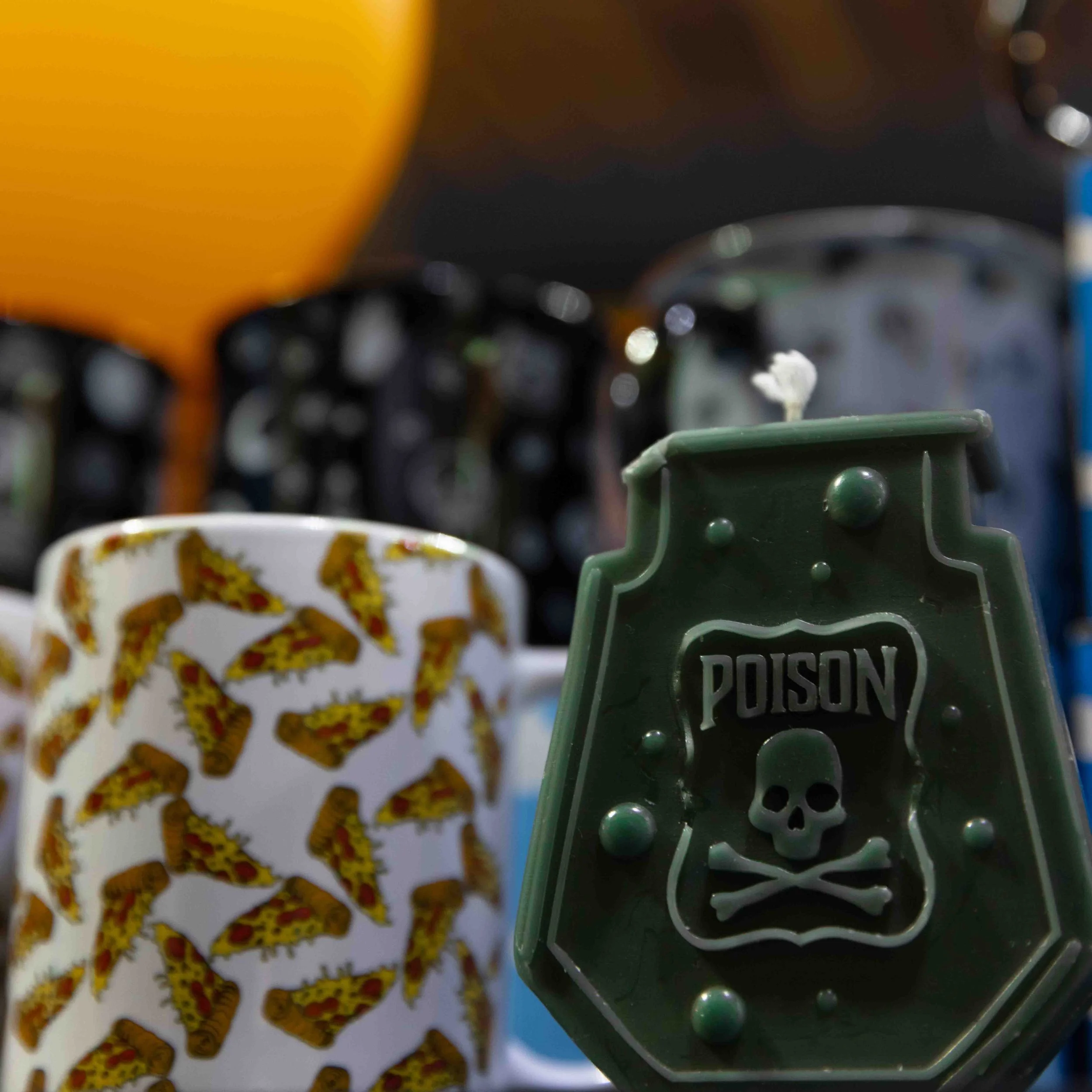 A green toy container with a skull and crossbones symbol and the word 'POISON' on it, placed next to a white mug decorated with pizza slices, and blurred shelves of cans in the background.