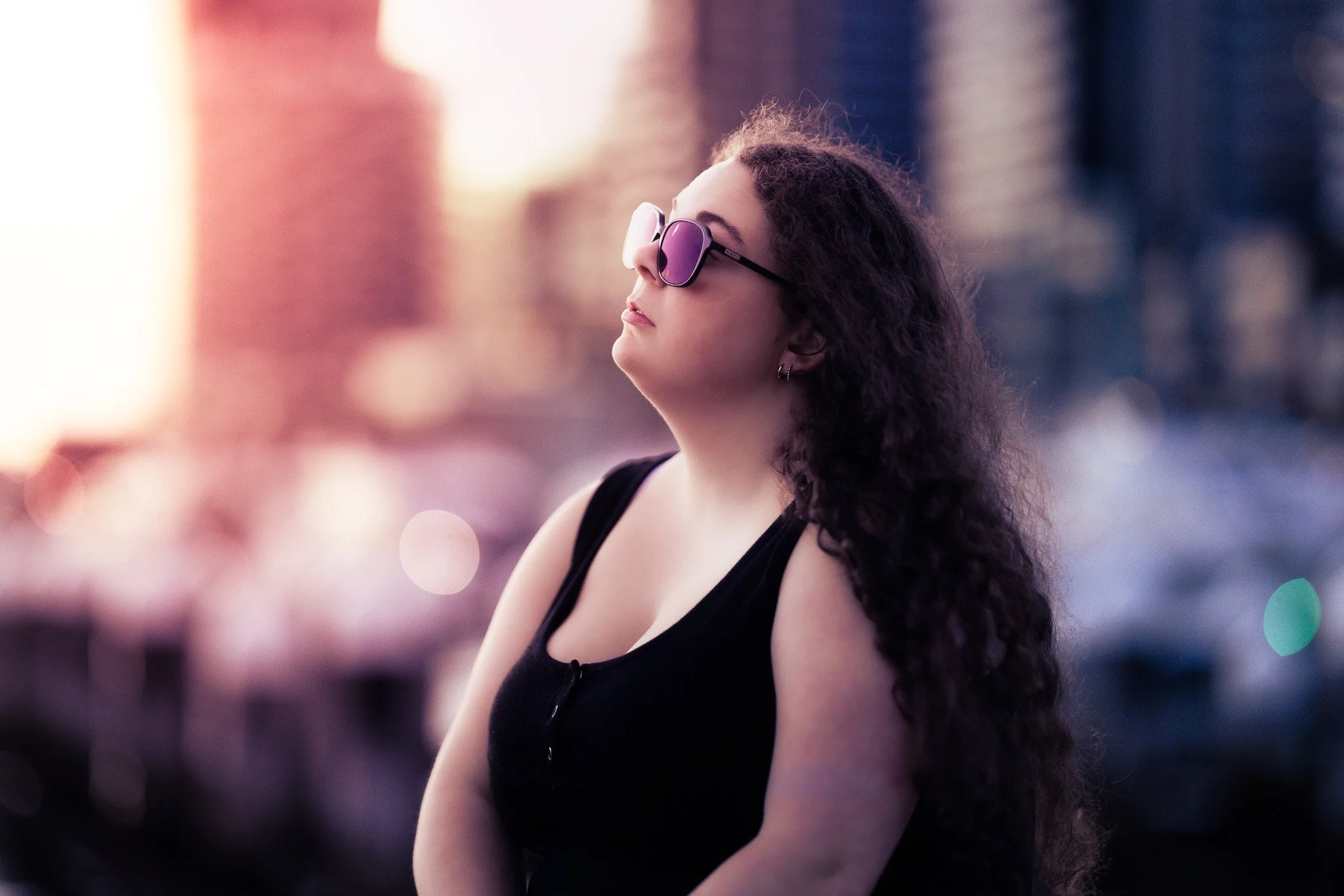 A woman with long, curly hair wearing sunglasses and a black sleeveless top, standing outdoors during sunset with a blurry cityscape background.