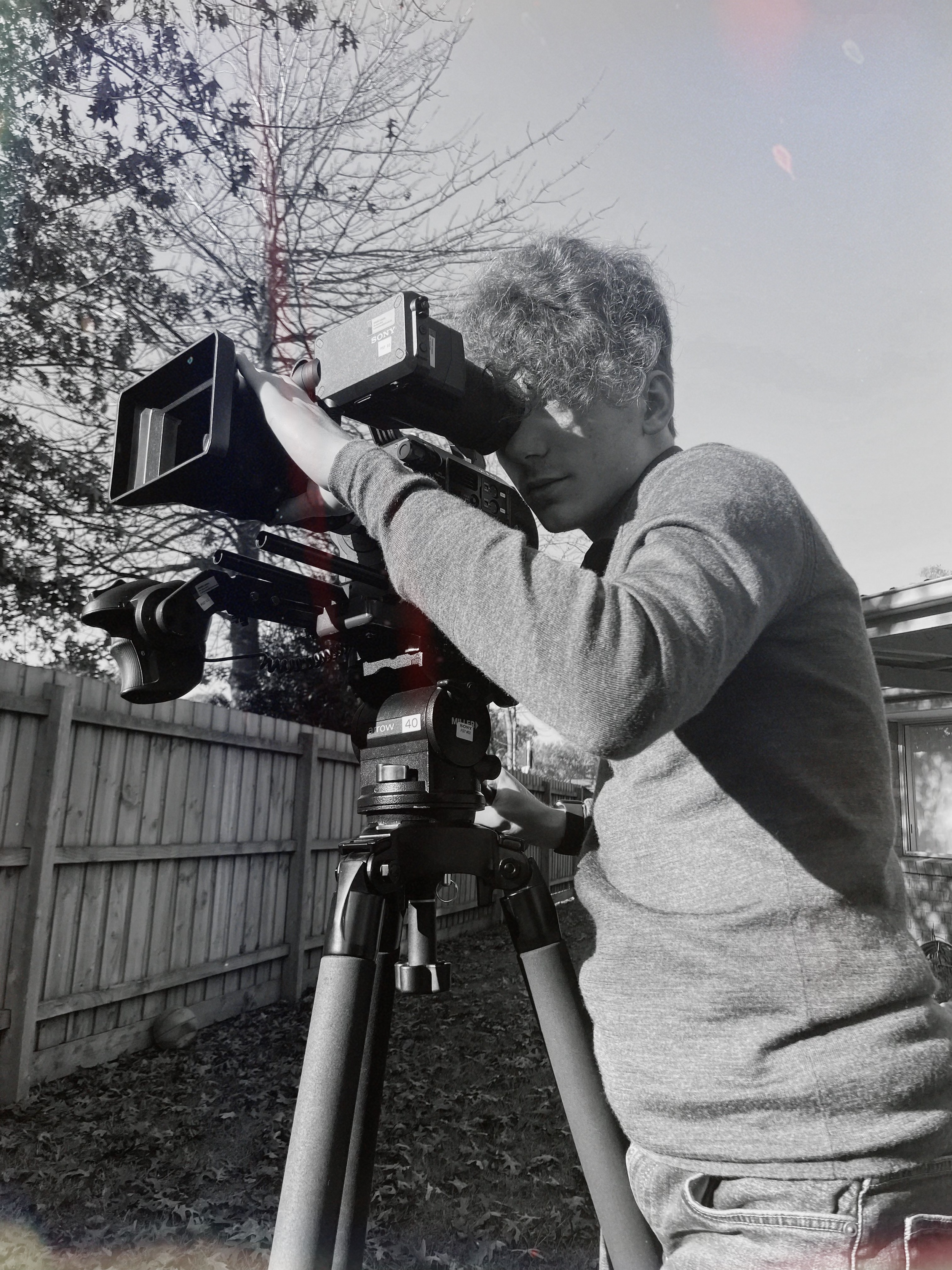 Toby, a young person with curly hair operating a professional video camera outdoors, with a wooden fence and trees in the background.