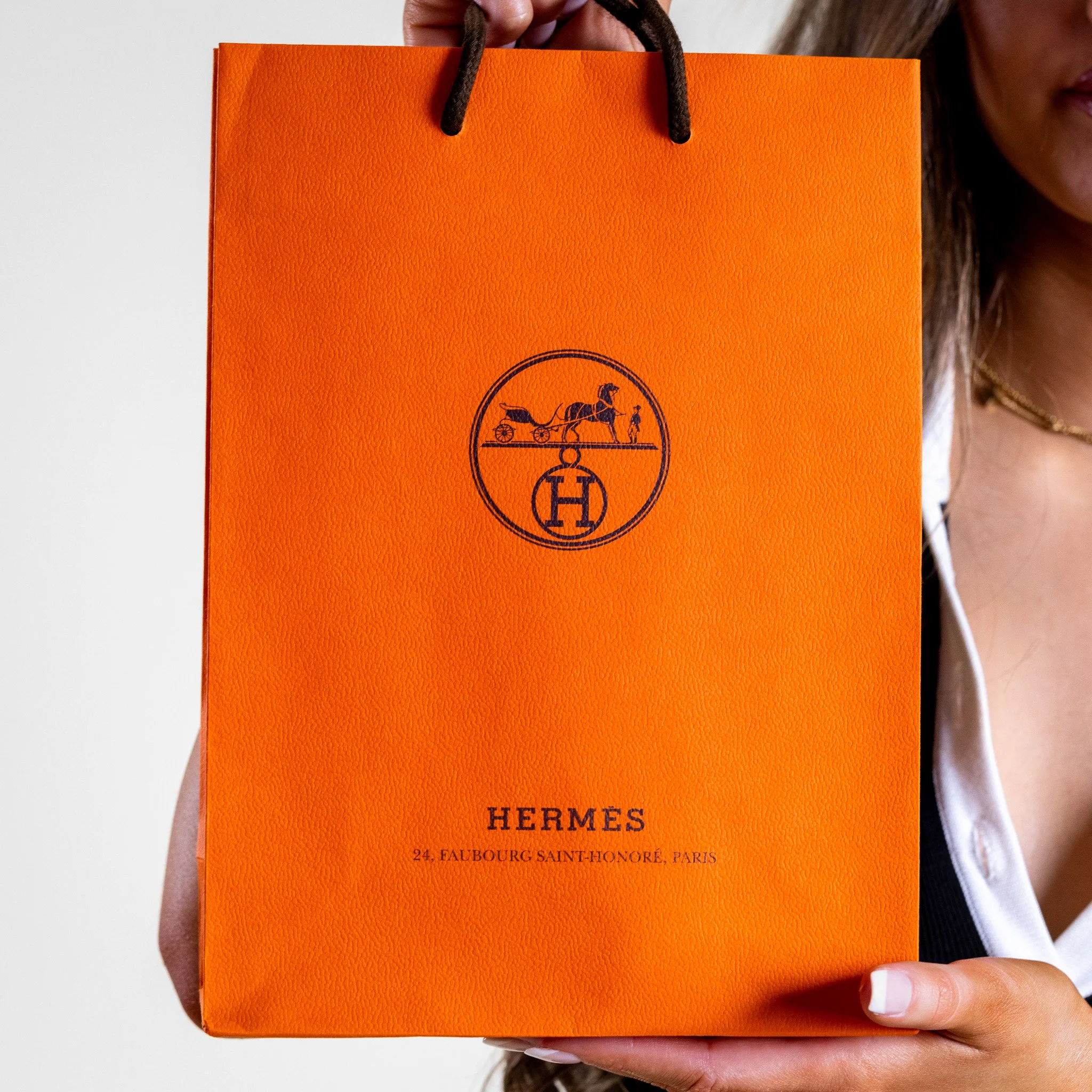 Person holding an orange Hermes shopping bag with black handles, featuring the Hermes logo and text indicating the store's address in Paris.