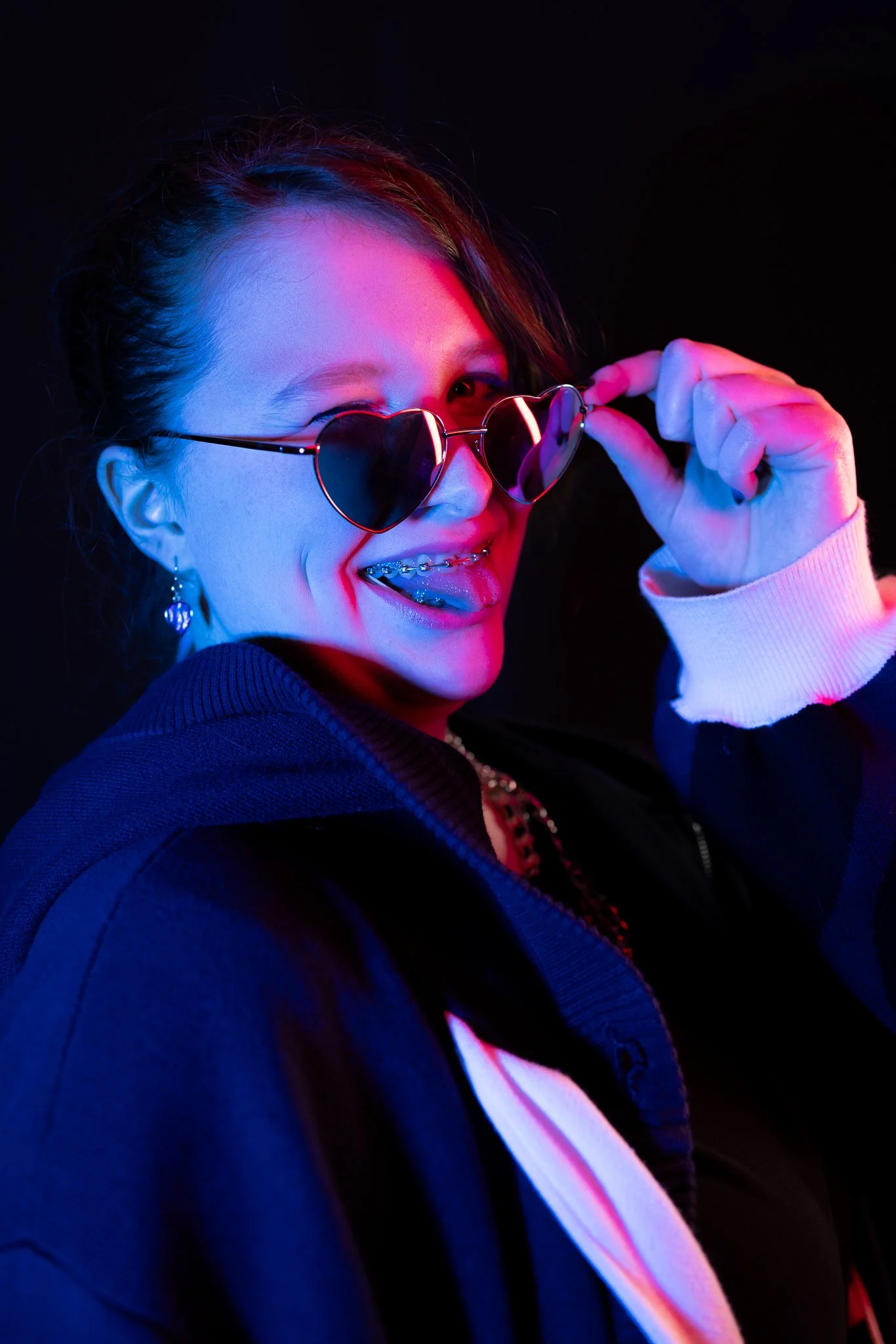 Young woman with braces wearing heart-shaped sunglasses, smiling, and holding the sunglasses with her right hand, against a dark background with pink and blue lighting.
