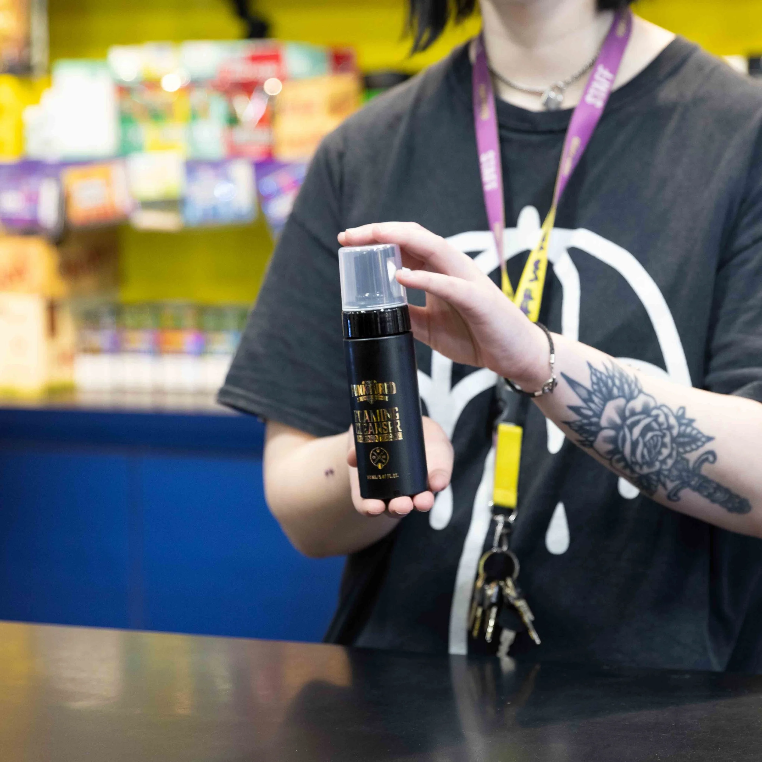 Person holding a black spray bottle with a spray top, with colorful shelves in the background.