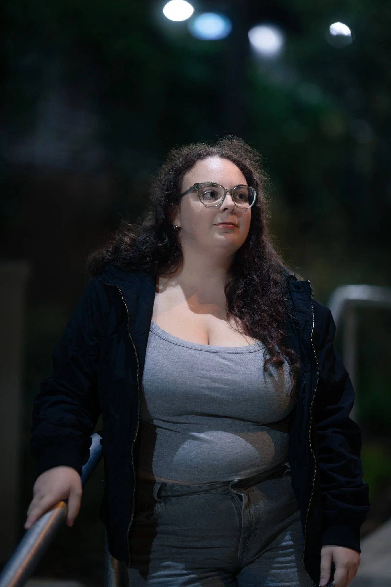A woman with curly dark hair, wearing glasses, a grey tank top, and a black jacket, standing outdoors at night near a metal railing.