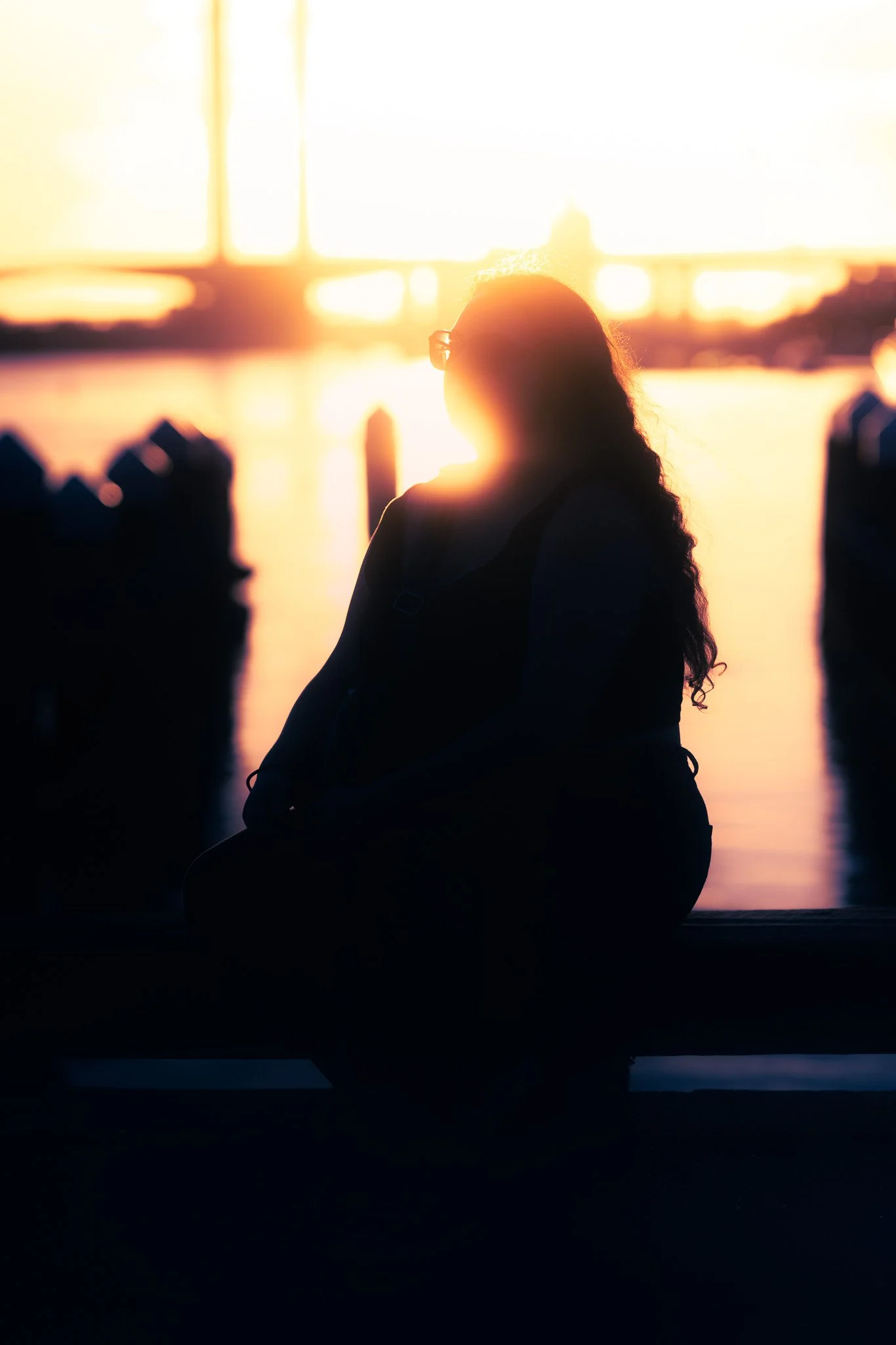 Silhouette of a person sitting by water during sunset with a bridge in the background.