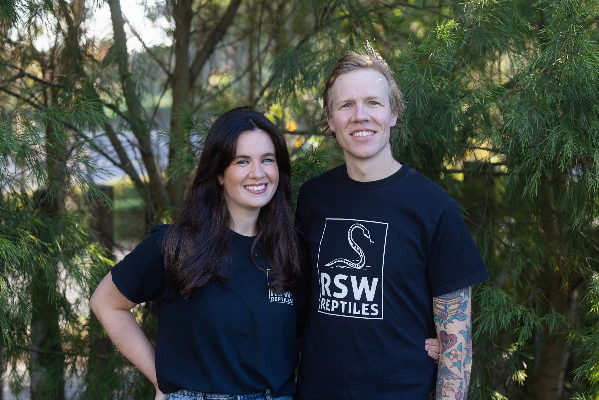 A smiling woman with long dark hair and a man with short light hair, both wearing black T-shirts with a reptile logo and 'RSW Reptiles' text, standing outdoors in front of green foliage.