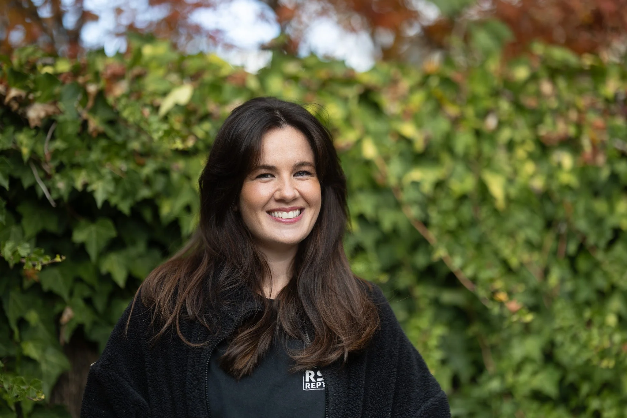 A smiling woman with long brown hair in front of green ivy and autumn trees.