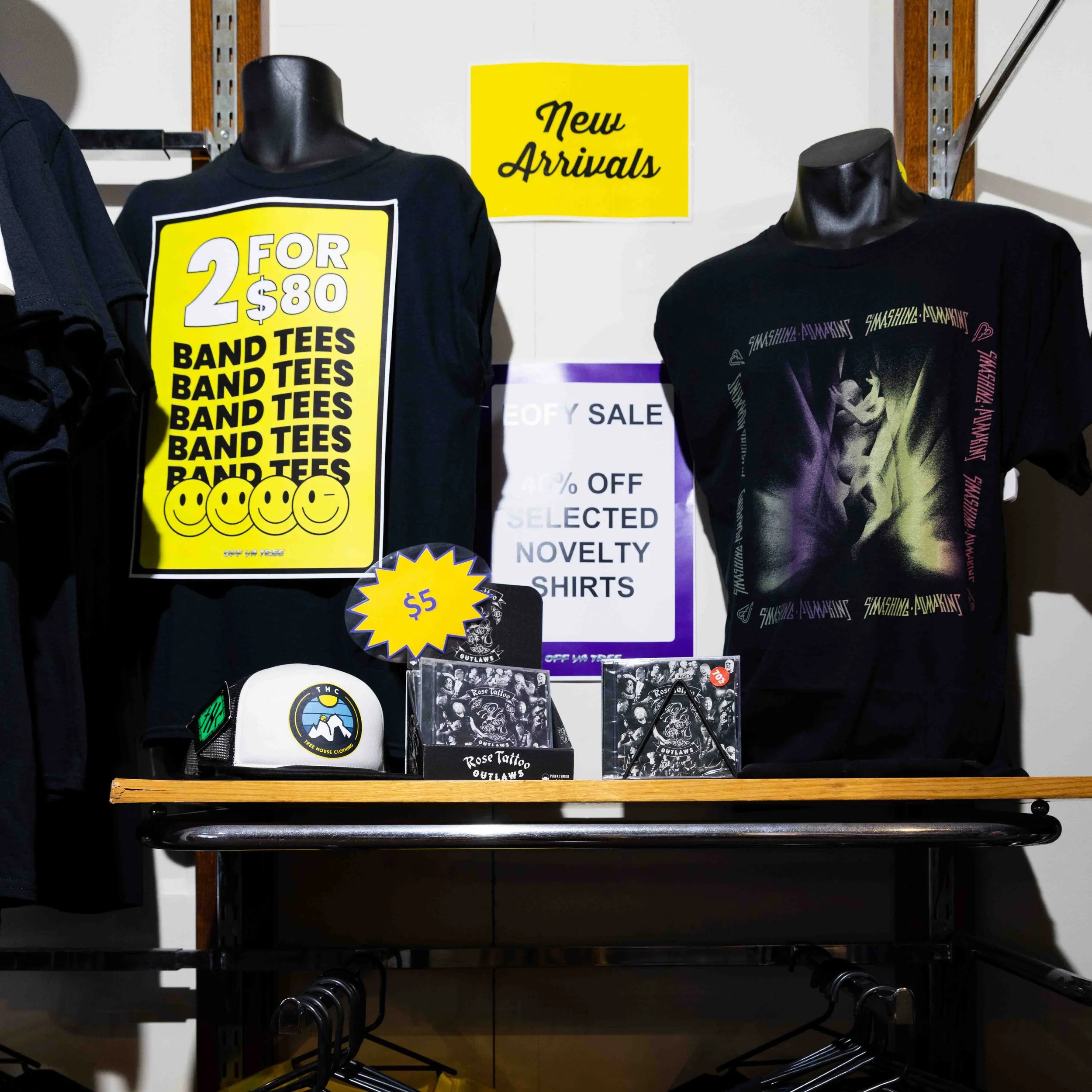 Clothing store display with black band t-shirts, one with a yellow sign advertising 2 for $80, and another with a graphic of a woman, with a yellow 'New Arrivals' sign above. There are also small items like a white cap with a mountain logo, rock tatt