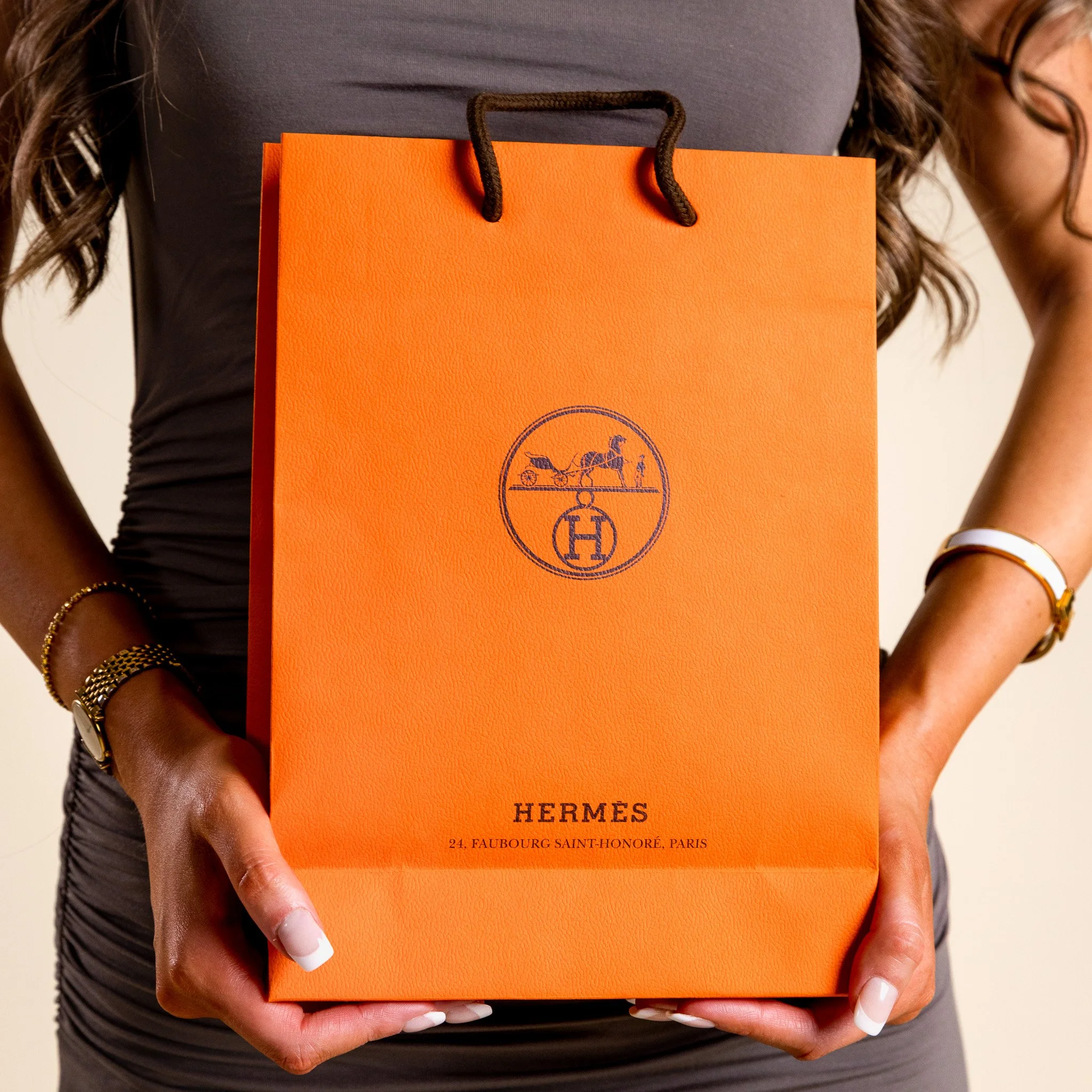 Person holding an orange Hermès shopping bag with black handles and a logo, wearing a dark gray dress and jewelry.