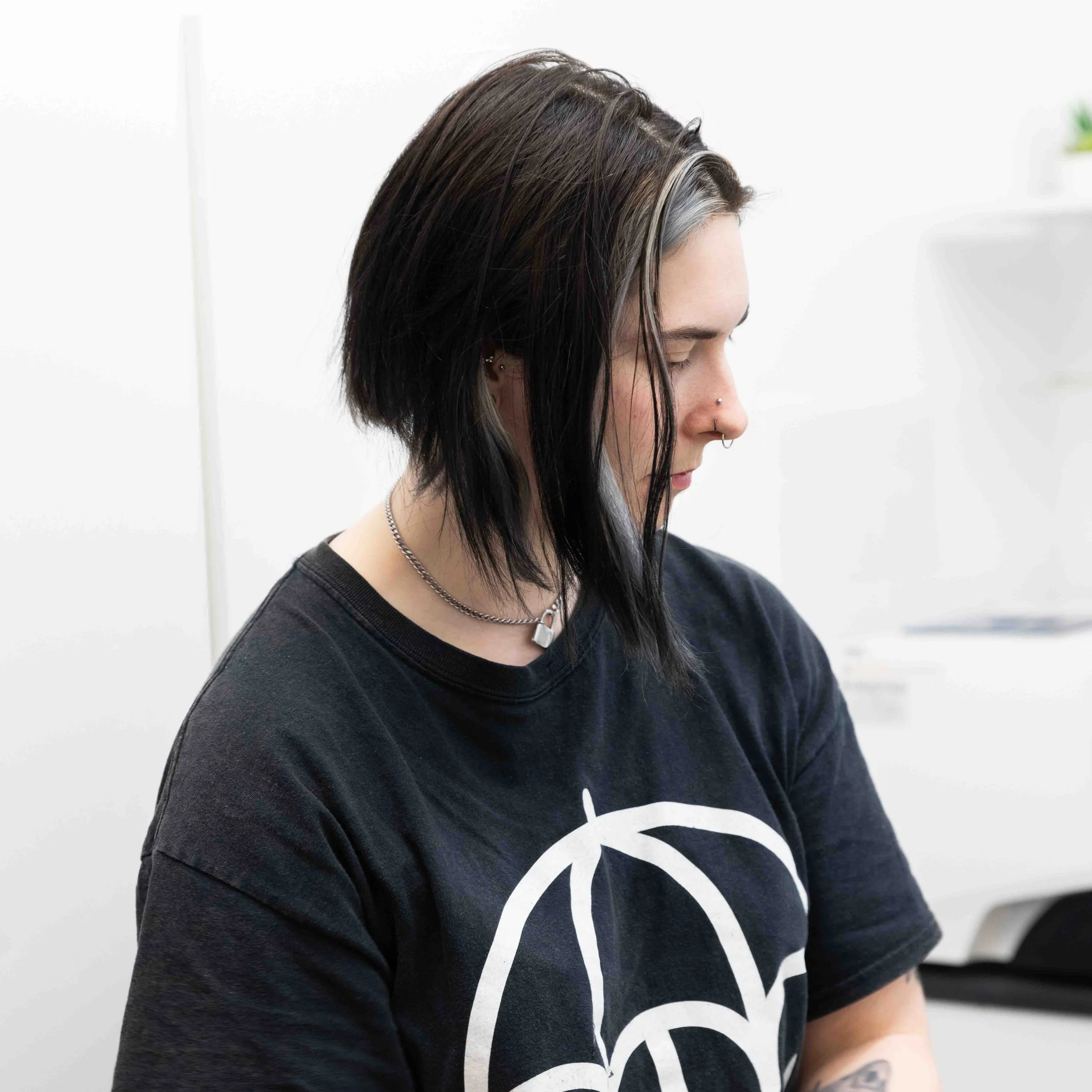 A person with black and gray hair, multiple facial piercings, and a silver necklace, looking down, wearing a black t-shirt with a white graphic design, standing in a bright indoor setting.