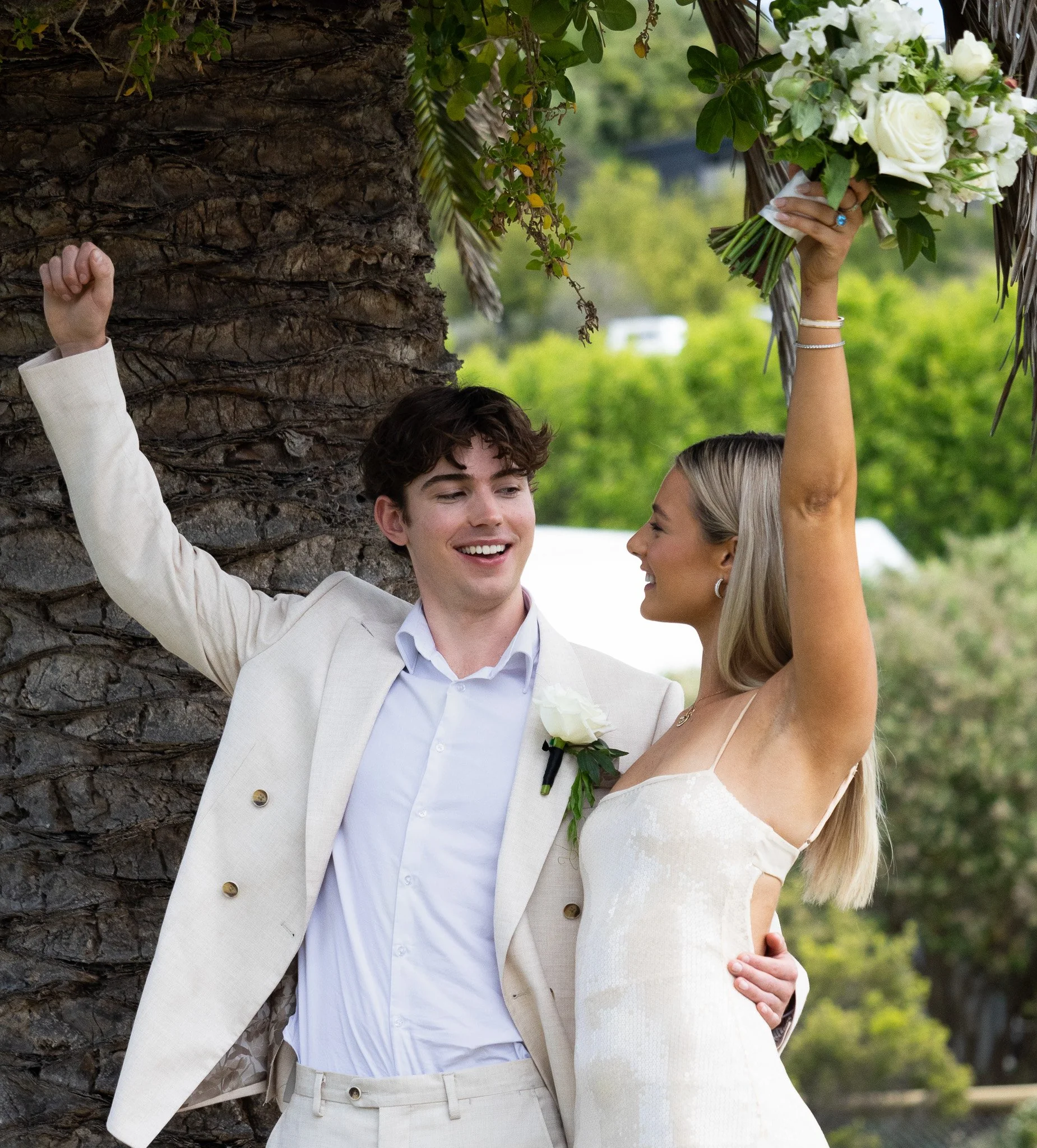 A smiling newlywed couple celebrating outdoors, with the woman raising a bouquet of white roses and greenery in the air. The man is dressed in a light suit with a boutonniere, and the woman is in a white dress, standing near a palm tree with lush gre