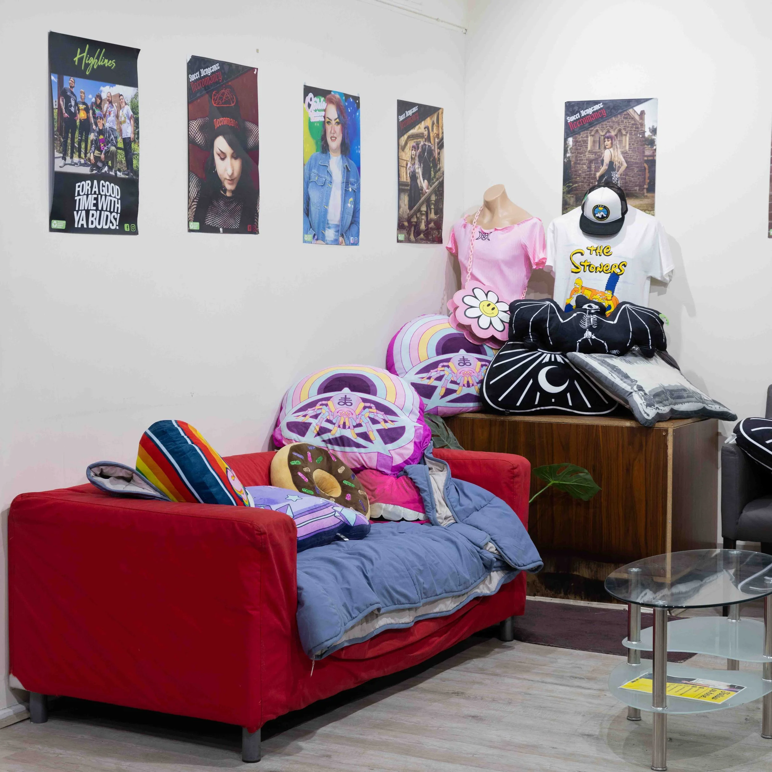 Living room decorated with posters on the wall, a red sofa with colorful pillows including a donut pillow, a blue blanket, and a padlock-shaped pillow, and a wooden display table with more pillows and stuffed toys. There are two mannequin torsos with