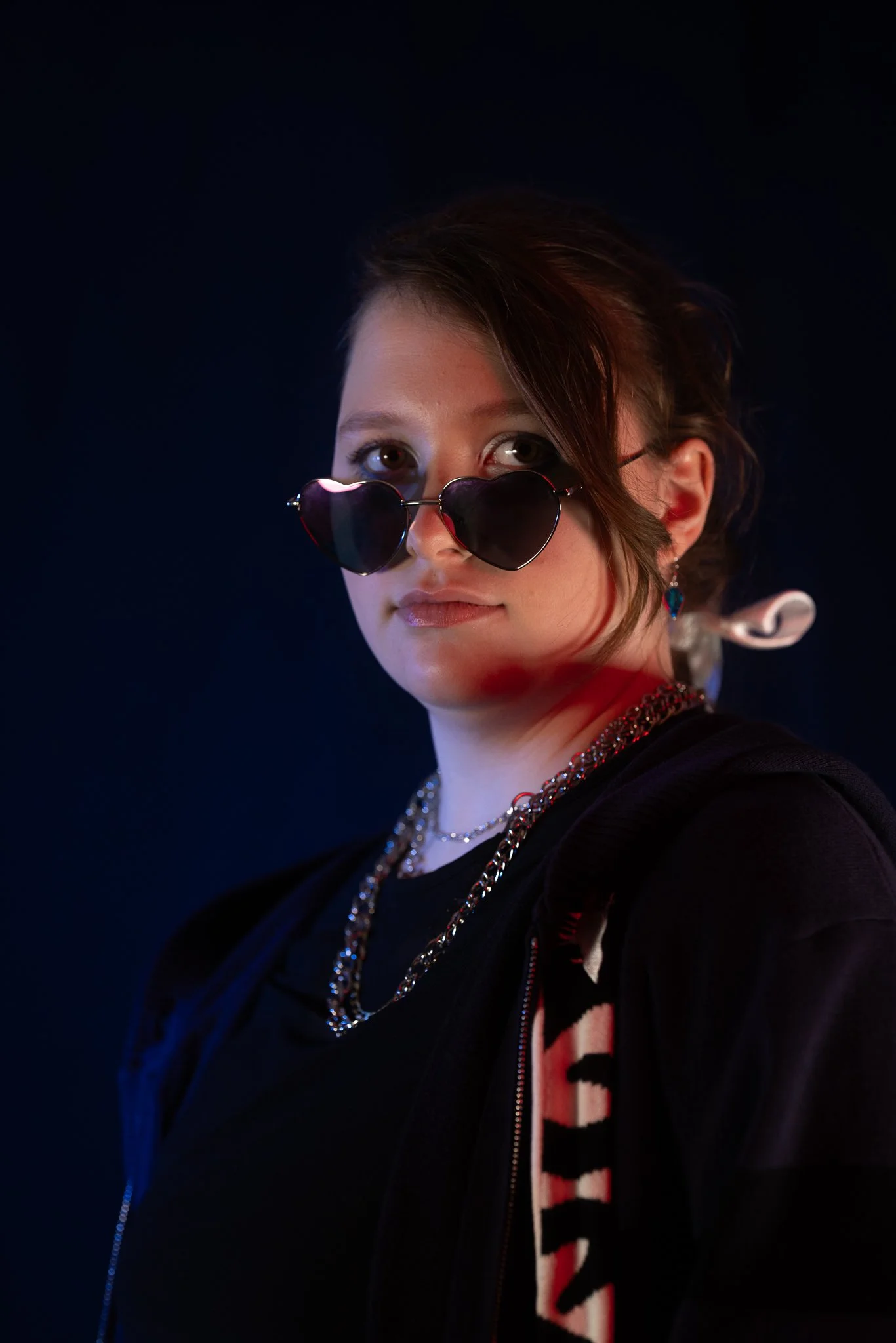 Young woman with short brown hair wearing heart-shaped sunglasses, layered necklaces, earrings, and a black hoodie against a dark background.